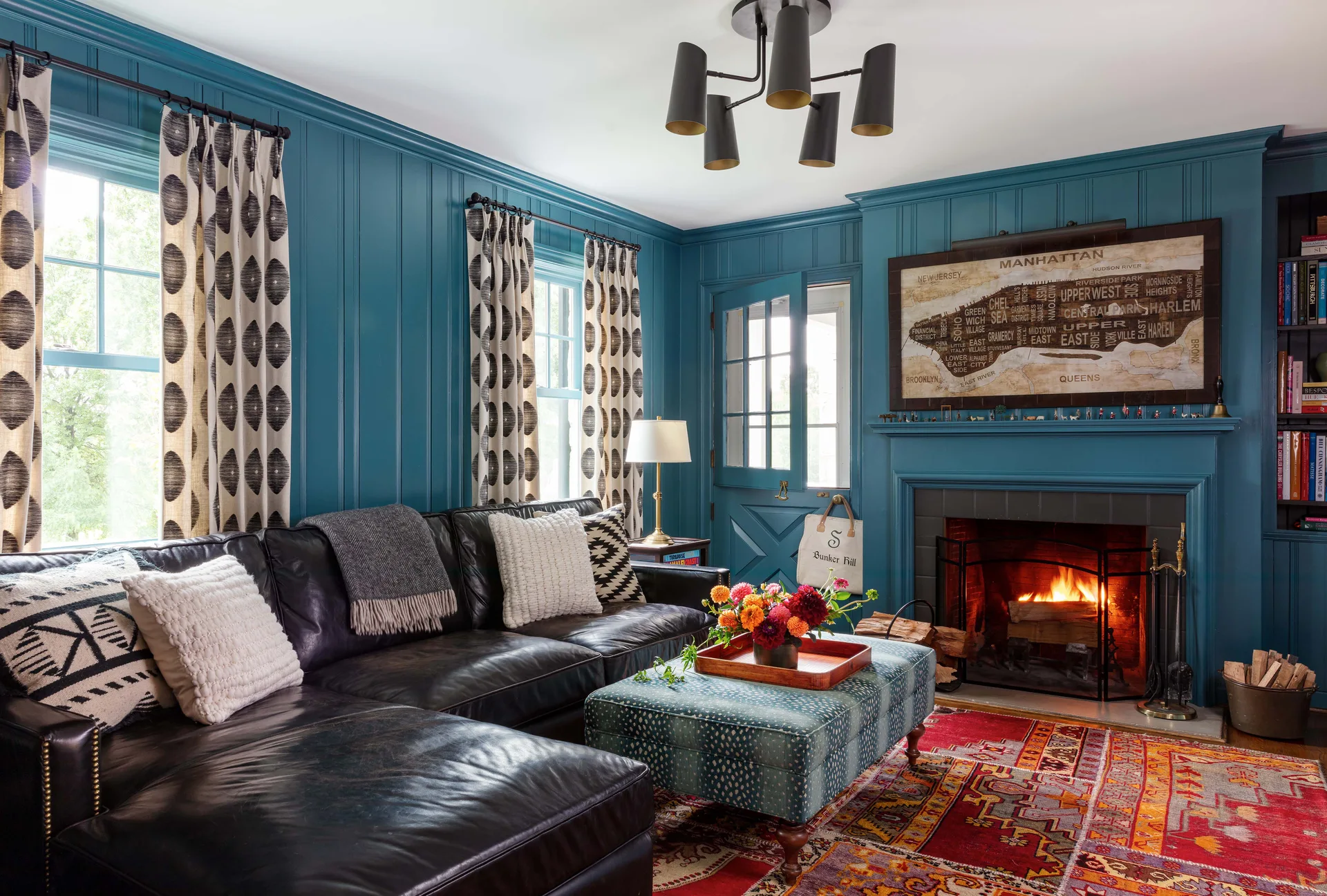 A cozy living room features deep teal wood-paneled walls, a black leather sectional sofa, a fireplace with a lit fire, and a colorful patterned area rug.