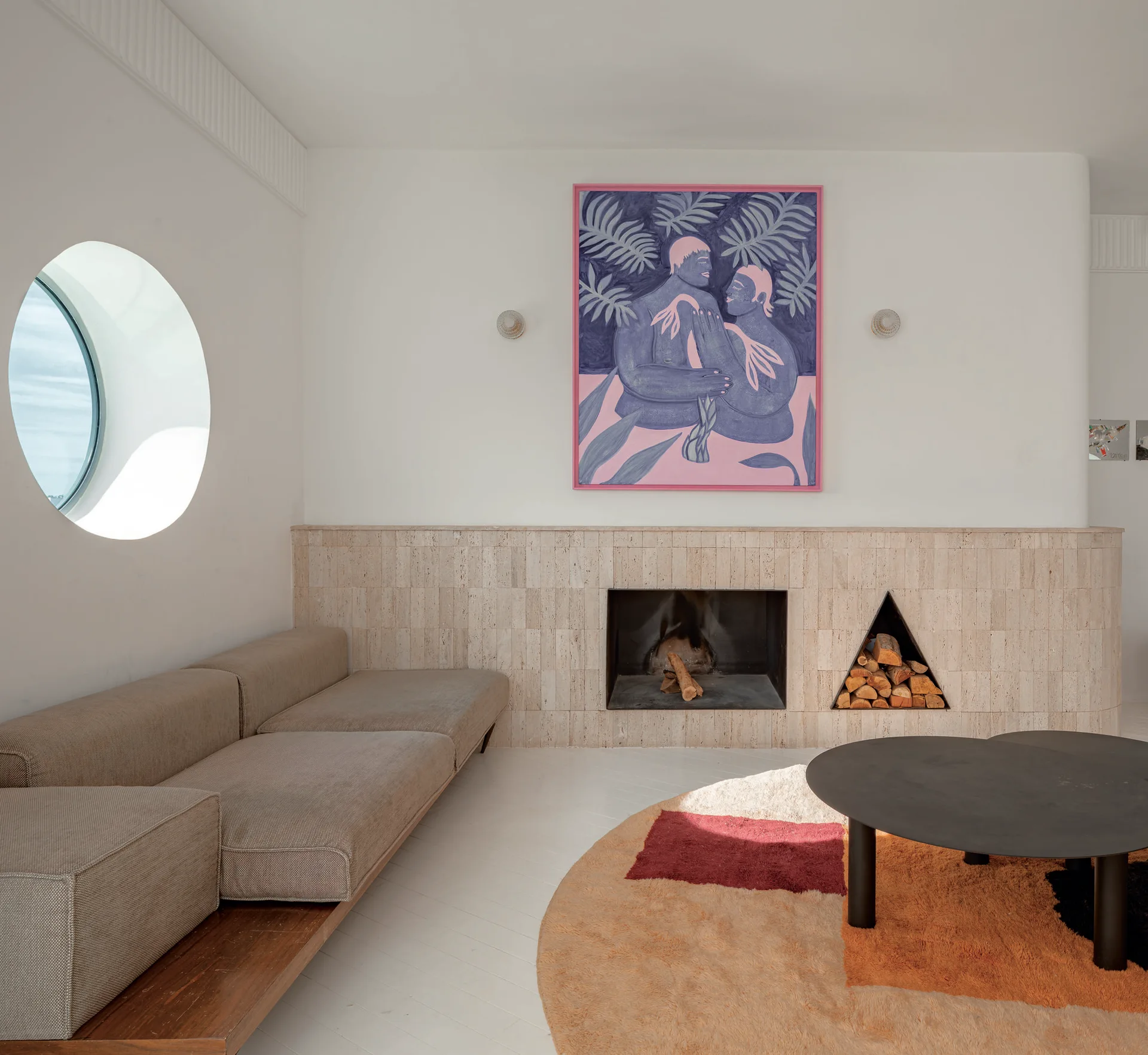 A minimalist living room featuring a low platform sofa, a circular window, and a travertine fireplace with a triangular log storage niche.