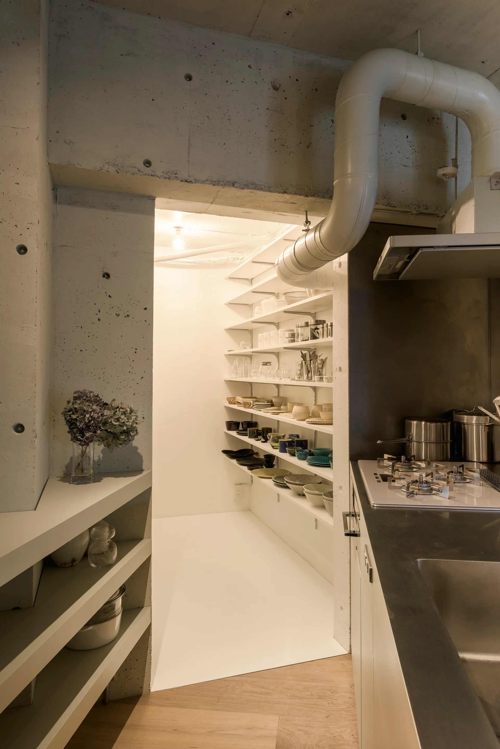 An industrial kitchen with exposed concrete walls, a stainless steel countertop, and a walk-in pantry filled with white open shelves.