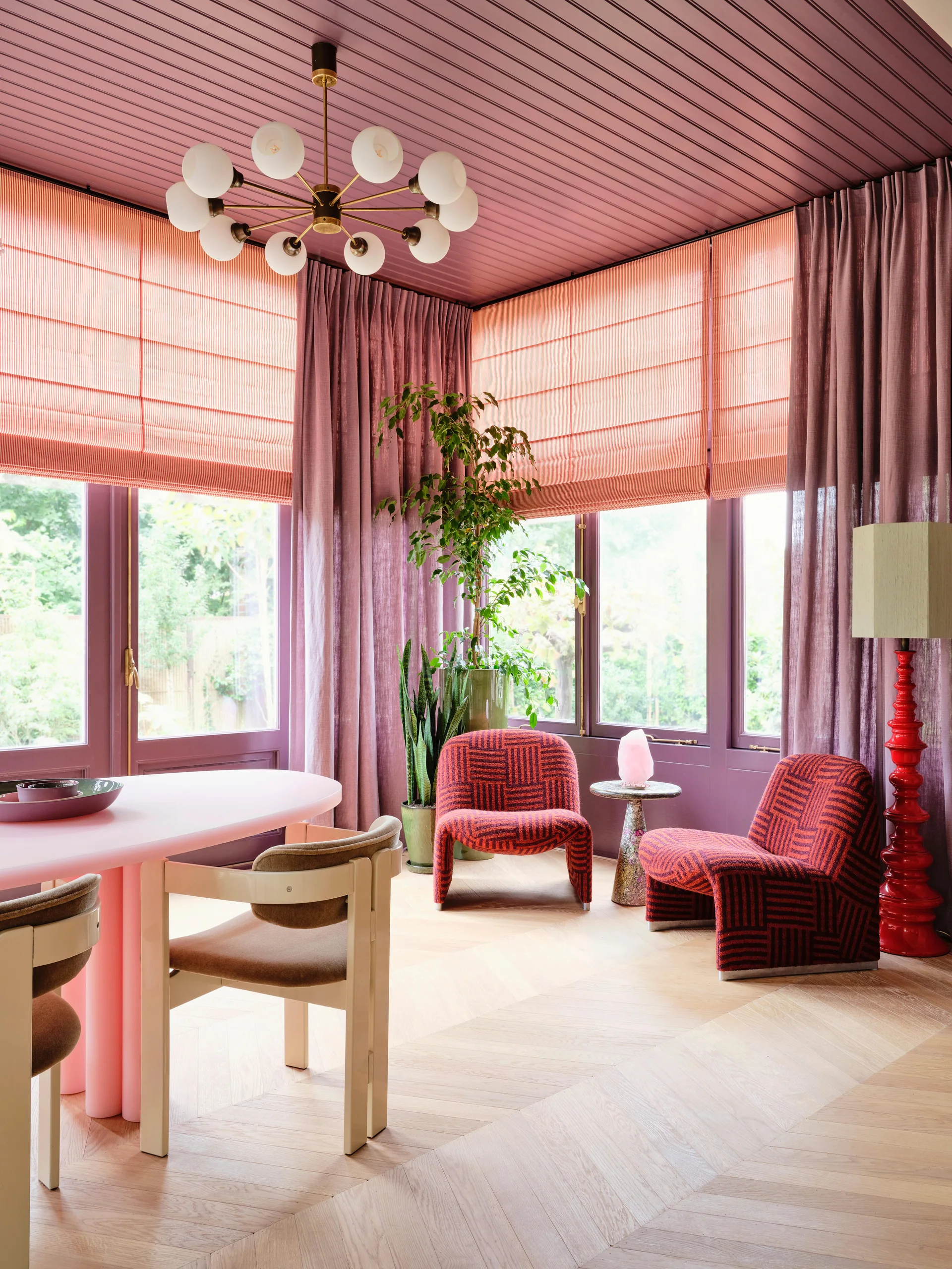 A vibrant dining space features a pink table, patterned red armchairs, and floor-to-ceiling windows framed by mauve curtains.