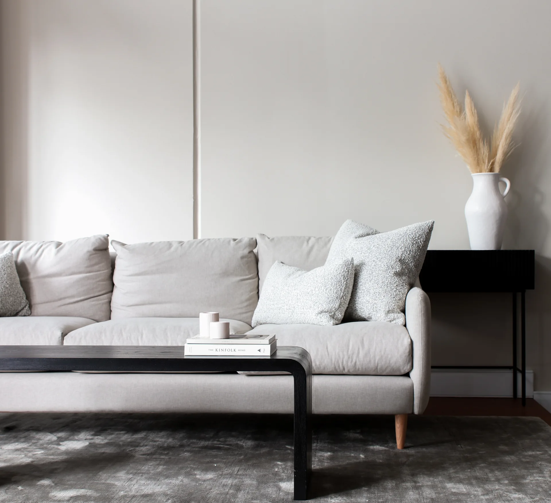 A light gray sofa with textured pillows sits behind a low black wood coffee table on a mottled gray rug in a minimalist living room.