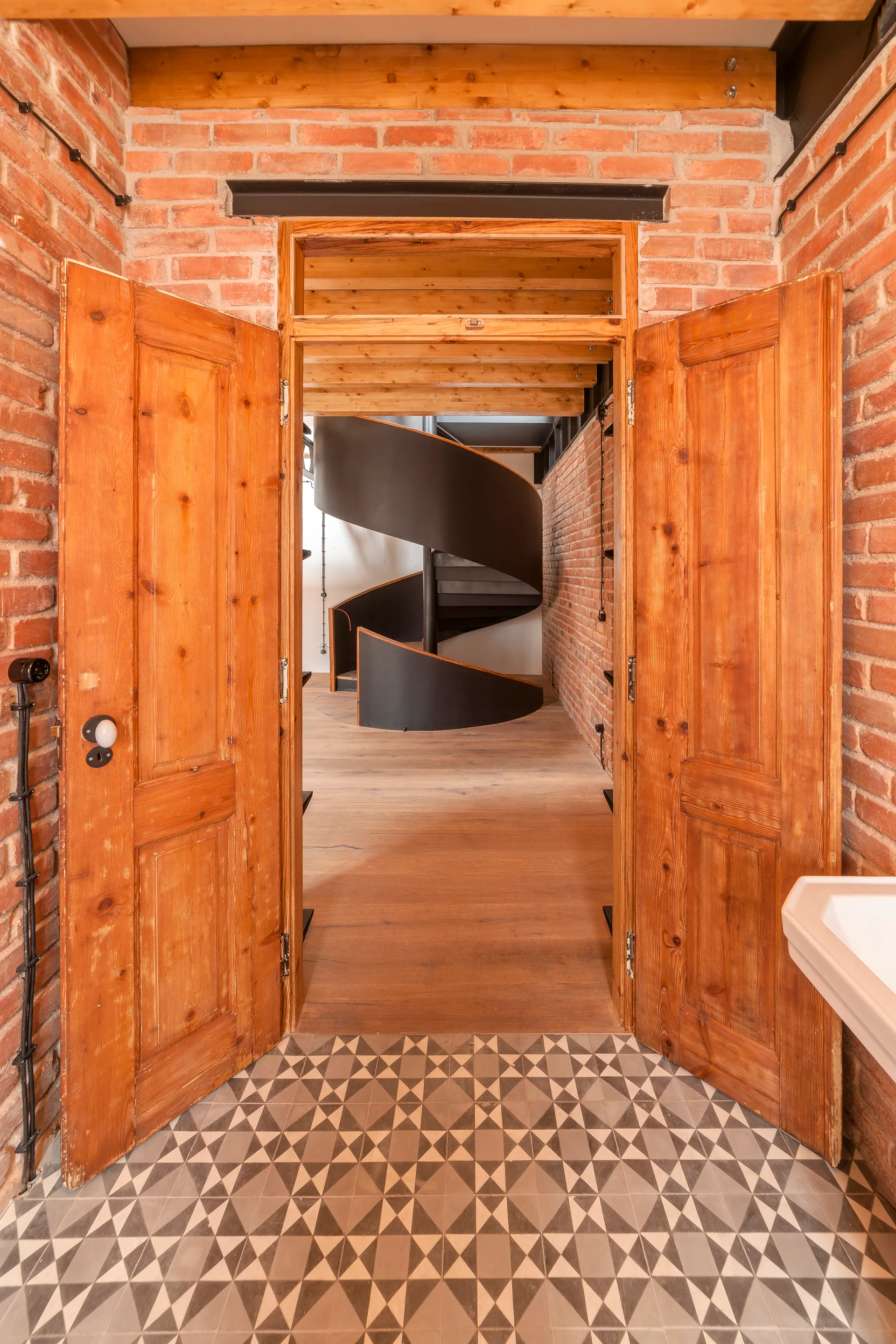 A view through open double wooden doors reveals a black metal spiral staircase, exposed red brick walls, and patterned geometric floor tiles.