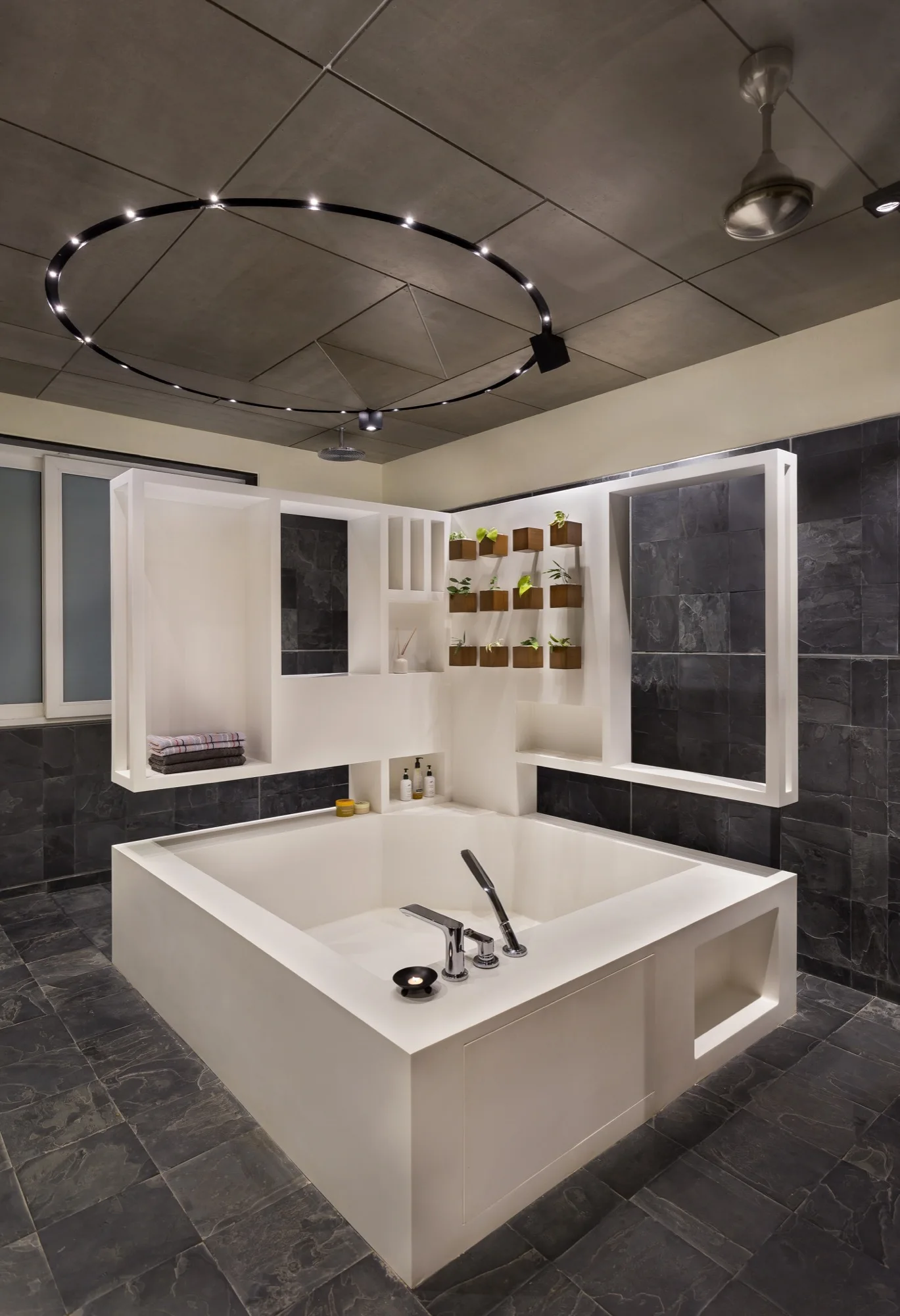 A square white soaking tub stands in a bathroom with dark grey stone tile walls, white geometric shelving, and a circular overhead light fixture.