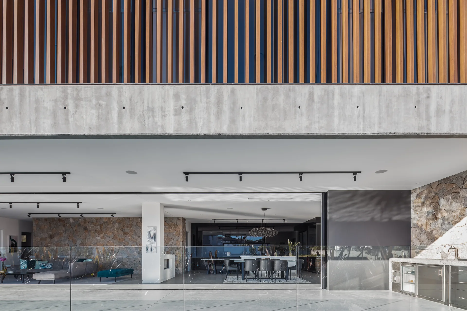 A contemporary patio features an exposed concrete beam, vertical wood slats, stone-clad walls, an outdoor kitchen, and a glass railing.