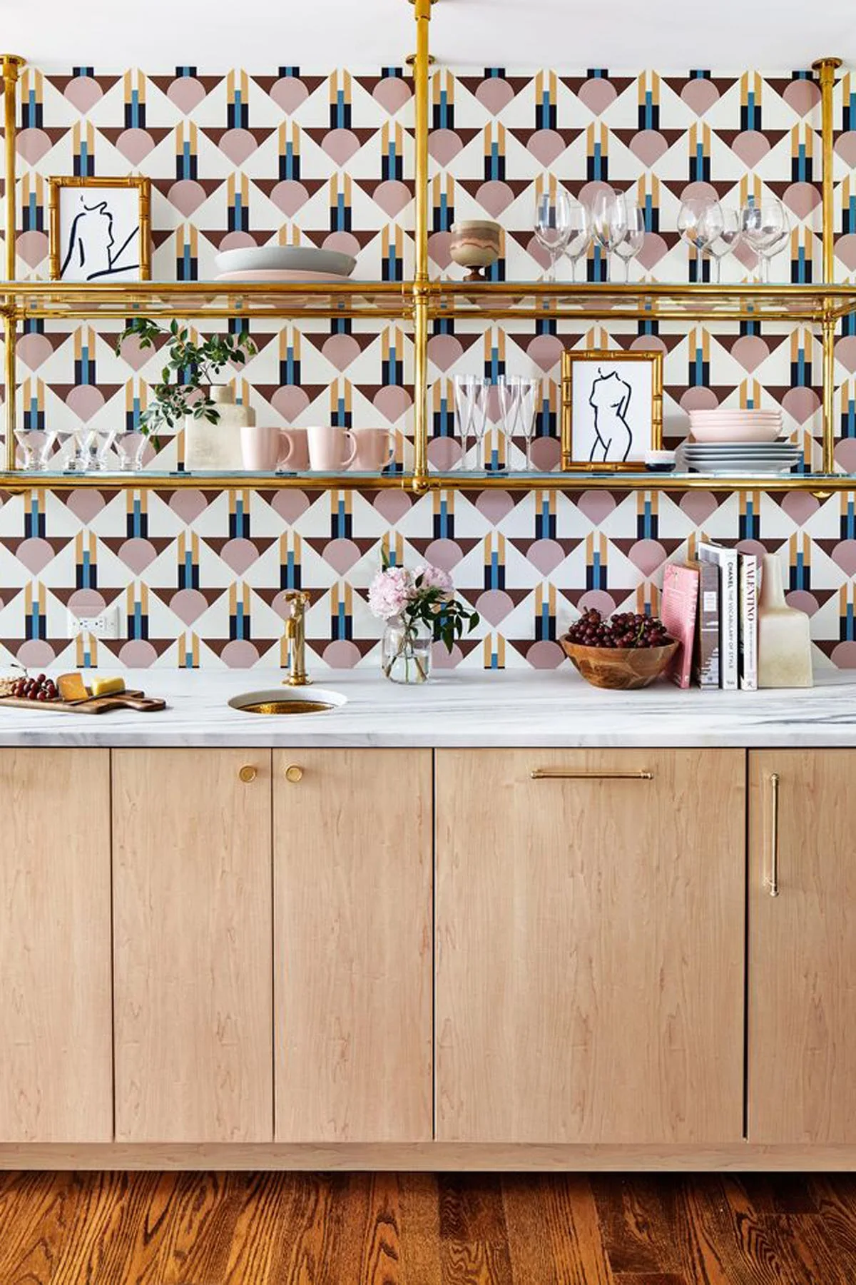 A modern home bar features light wood cabinets, a white marble countertop, brass open shelving, and pink geometric patterned wallpaper.