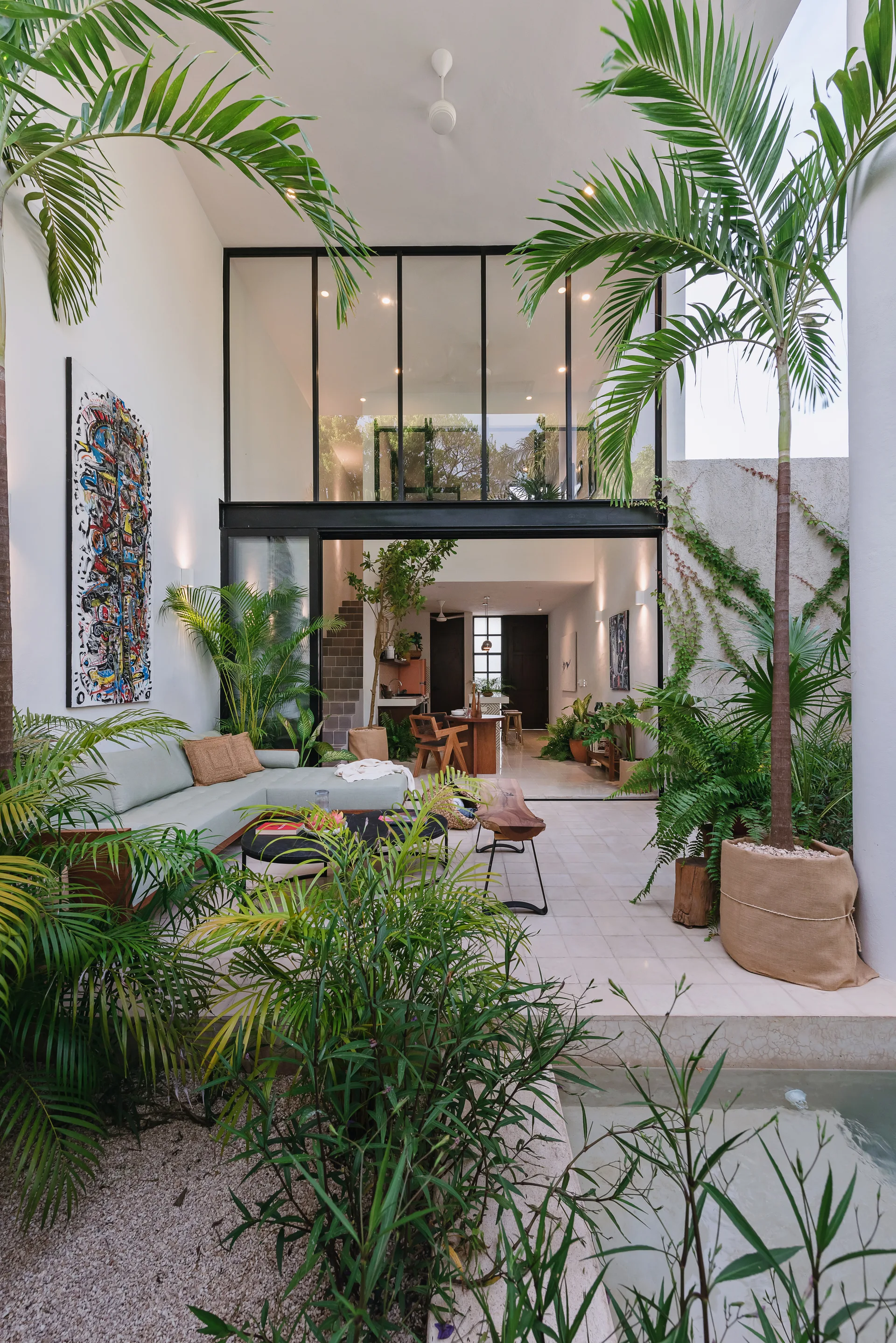 A contemporary outdoor patio features a light sectional sofa, lush potted palms in burlap bags, and a large black-framed glass wall leading inside.
