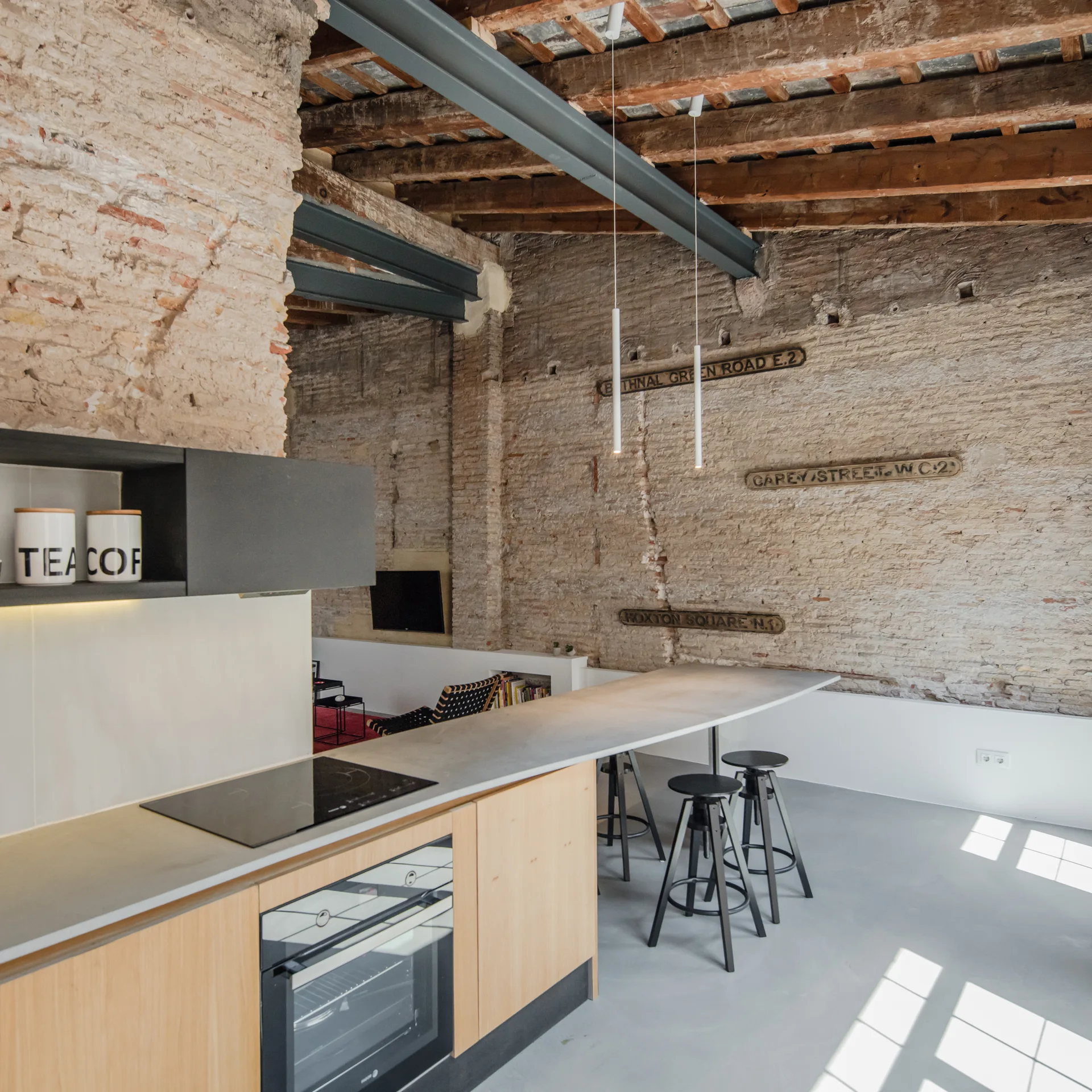 An industrial loft kitchen featuring a long grey concrete-style island, light wood cabinets, black bar stools, and an exposed brick wall.
