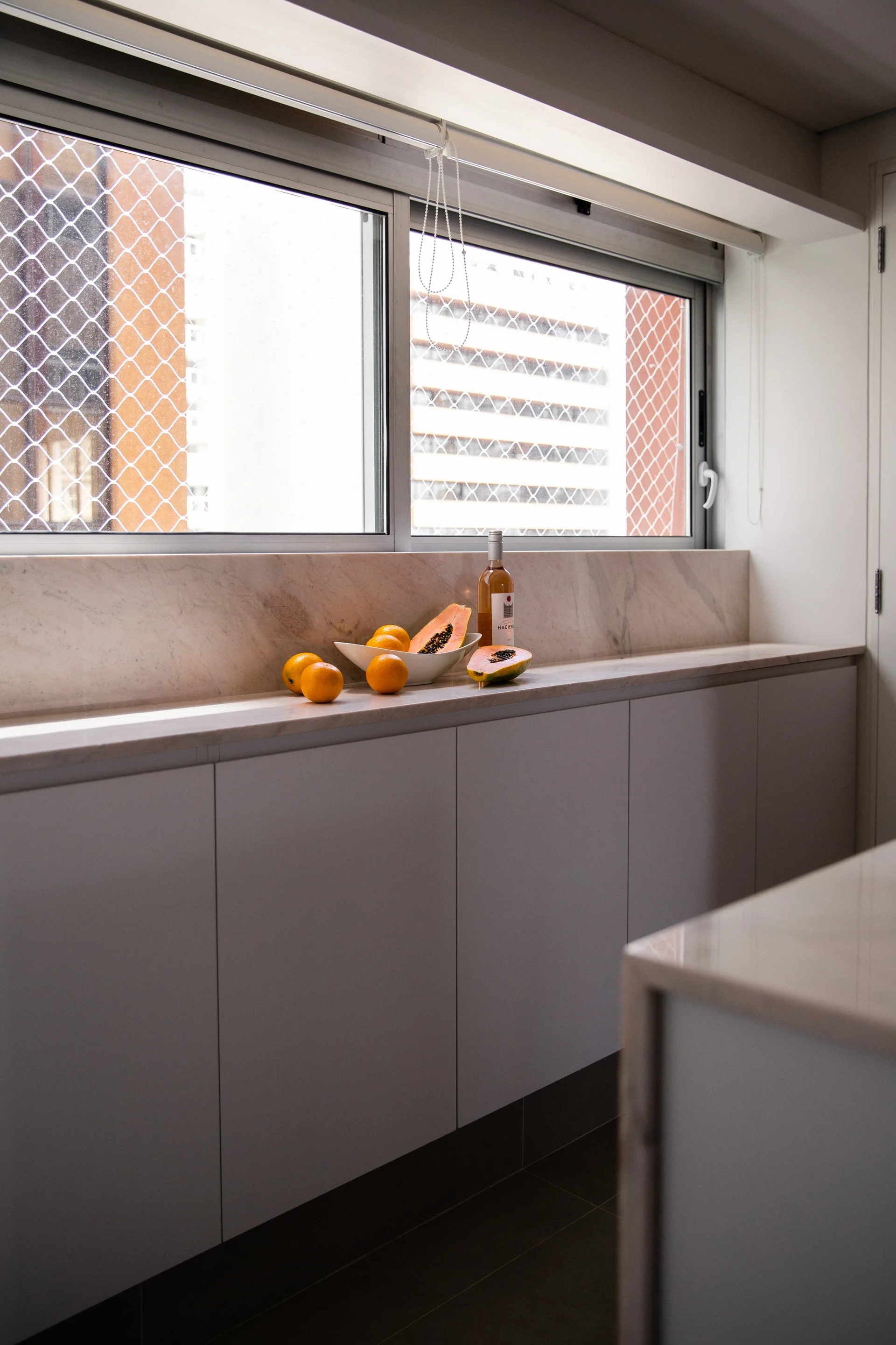 Beige flat-front kitchen cabinetry sits beneath a neutral stone countertop and low backsplash under a large sliding window with a safety screen.