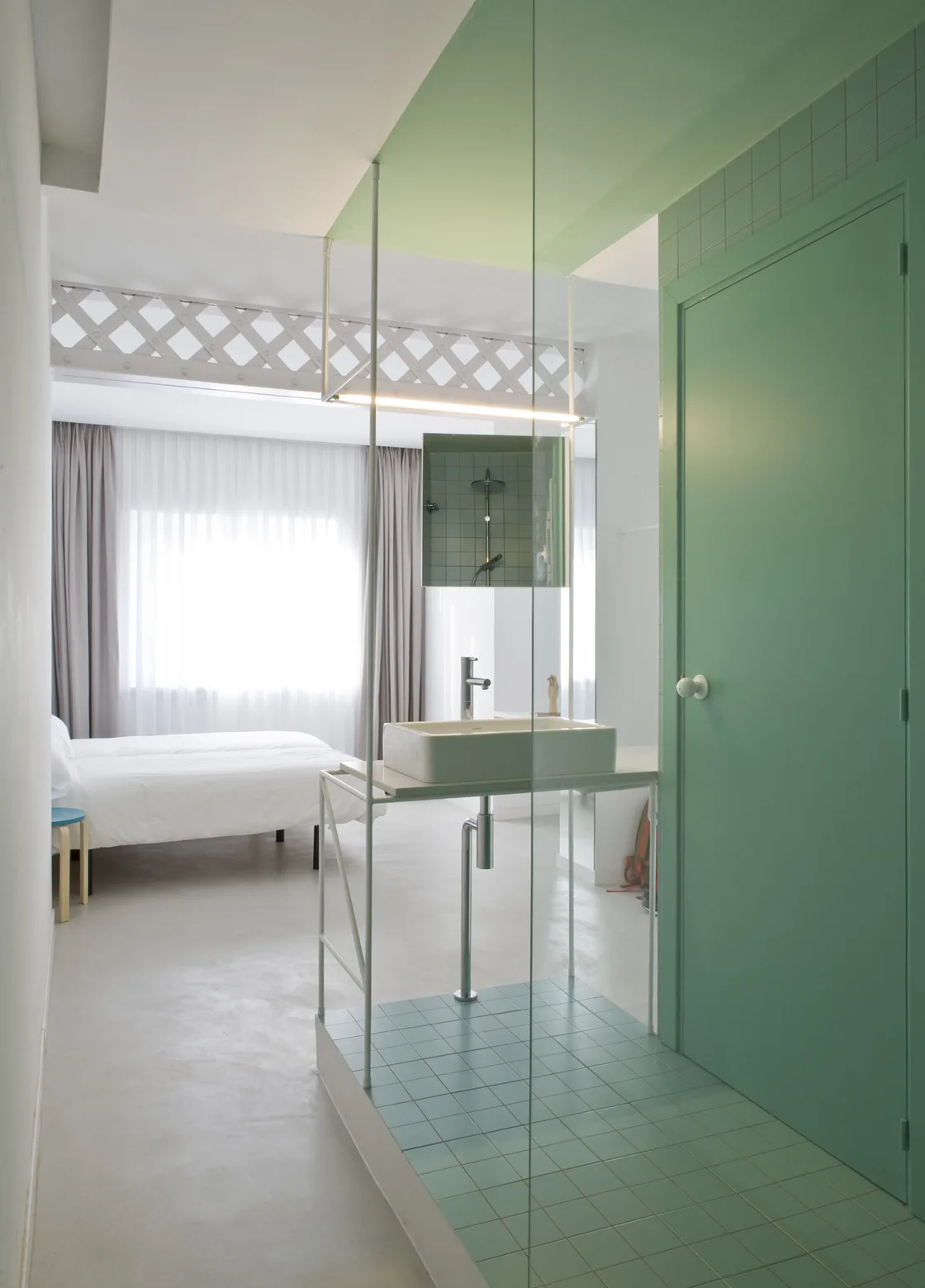 An open-concept bedroom featuring a white washbasin on a thin metal frame, a mint-green tiled platform, a green door, and a bed in the background.