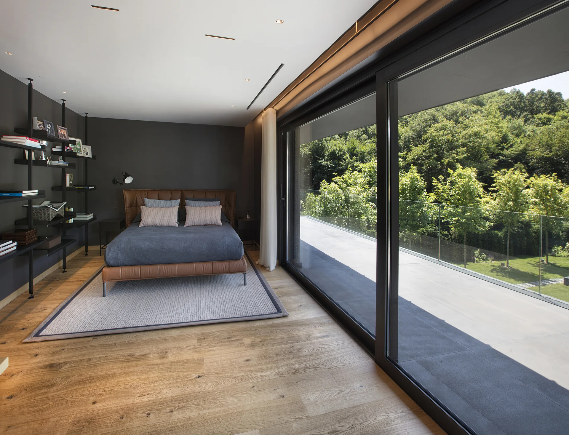 A modern bedroom featuring a brown leather bed, dark grey walls with black metal shelving, and sliding glass doors opening to a terrace.