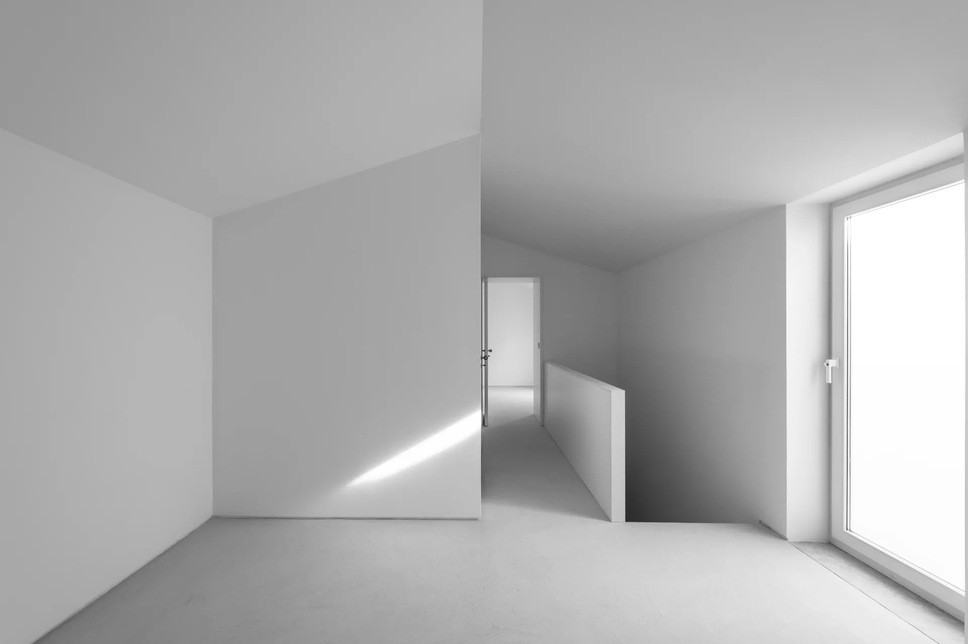 A minimalist white stairwell landing features smooth plaster walls, a sloped ceiling, and a large window that floods the space with bright light.