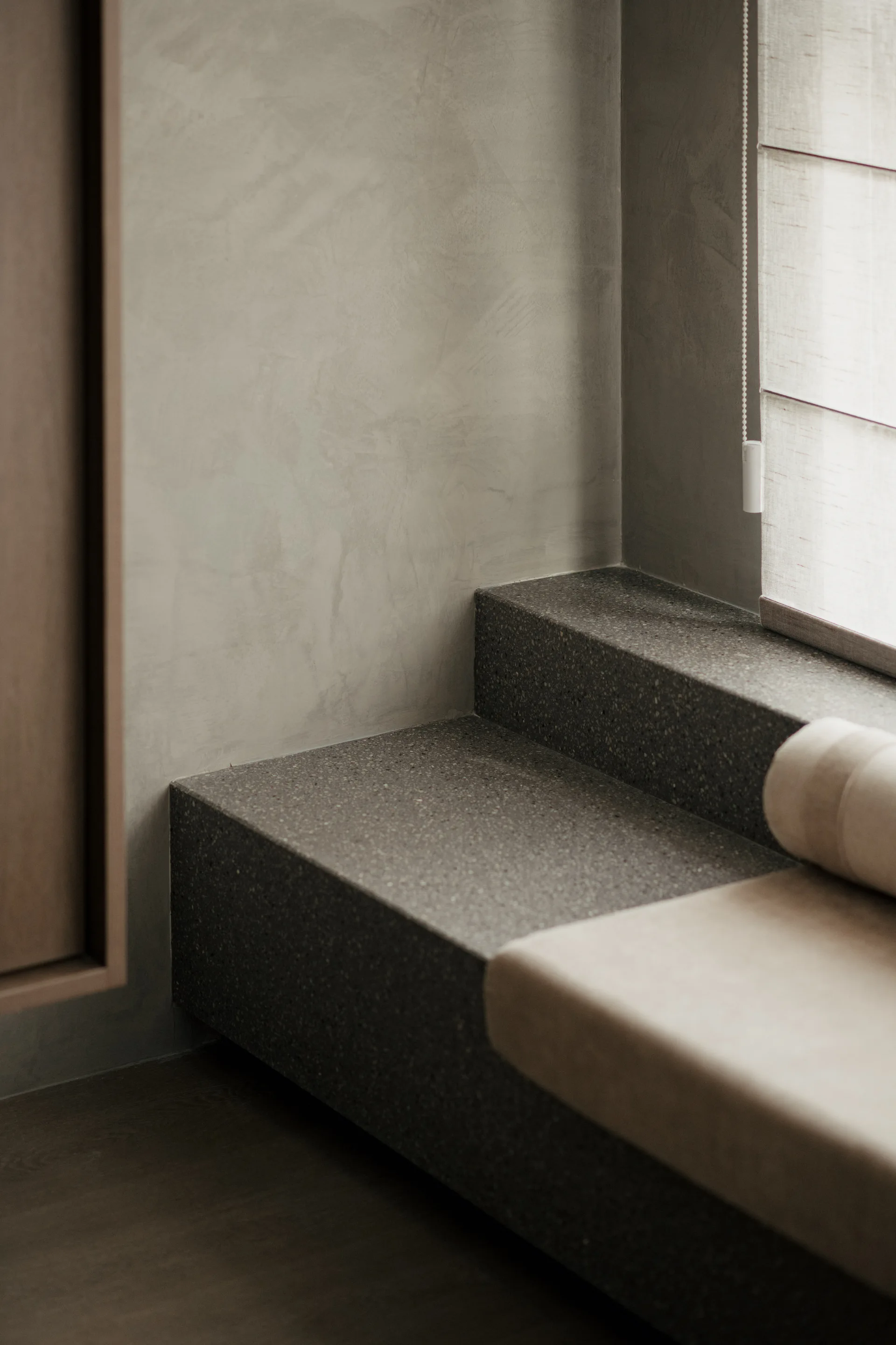 A close-up of a built-in stone window bench with a light beige cushion set against textured gray plaster walls.