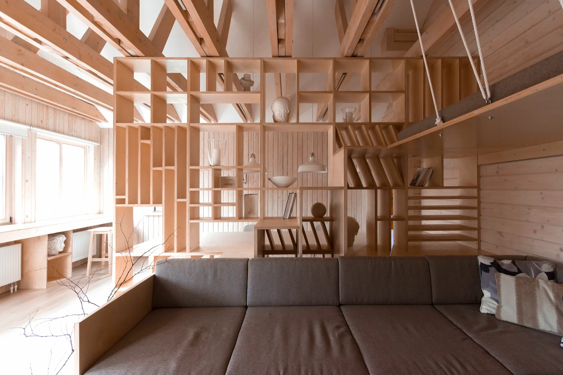 A living room features an open-grid wooden partition beneath exposed structural ceiling beams, with a large neutral sofa in the foreground.