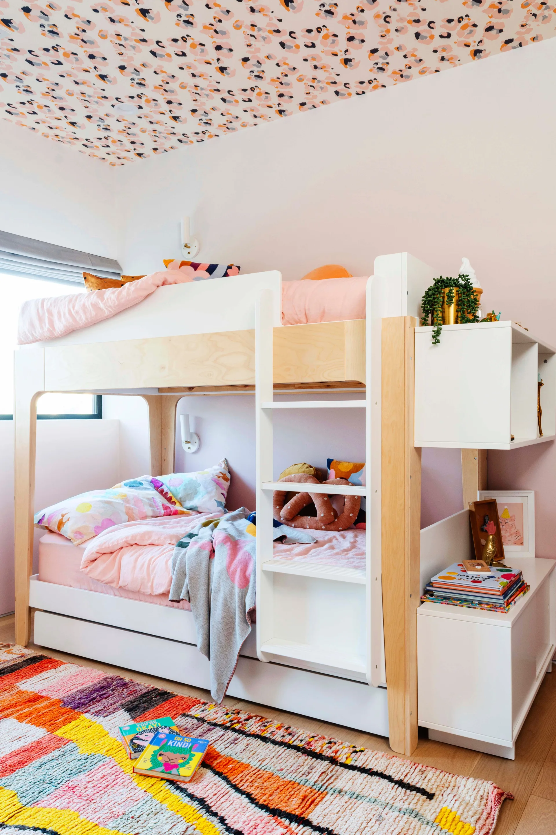 A modern wood and white bunk bed stands in a kids' bedroom beneath a spotted pattern ceiling, with a colorful striped rug on the floor.