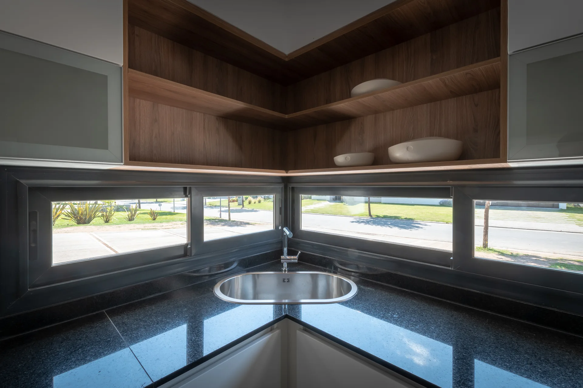A corner kitchen sink featuring a stainless steel basin set in a dark polished countertop below wraparound windows and L-shaped open wood shelving.