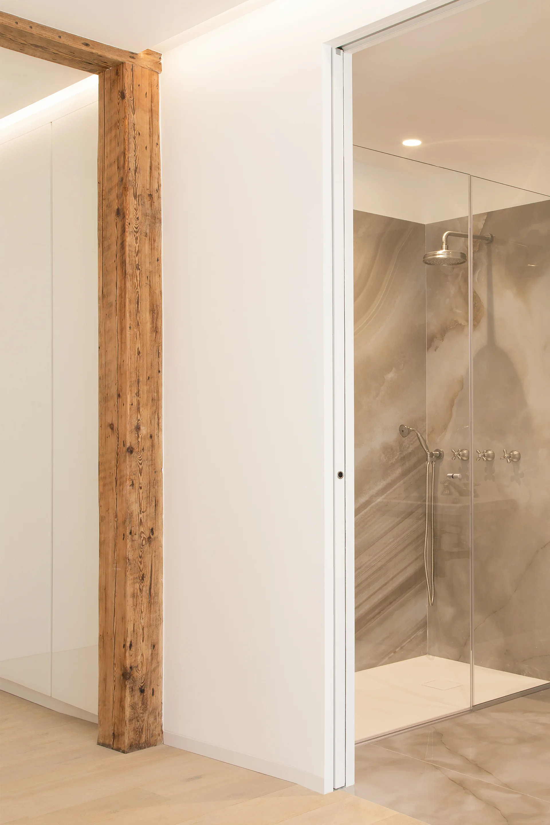 A modern bathroom featuring an exposed wooden post next to a glass-enclosed walk-in shower with beige marble-patterned tile walls.