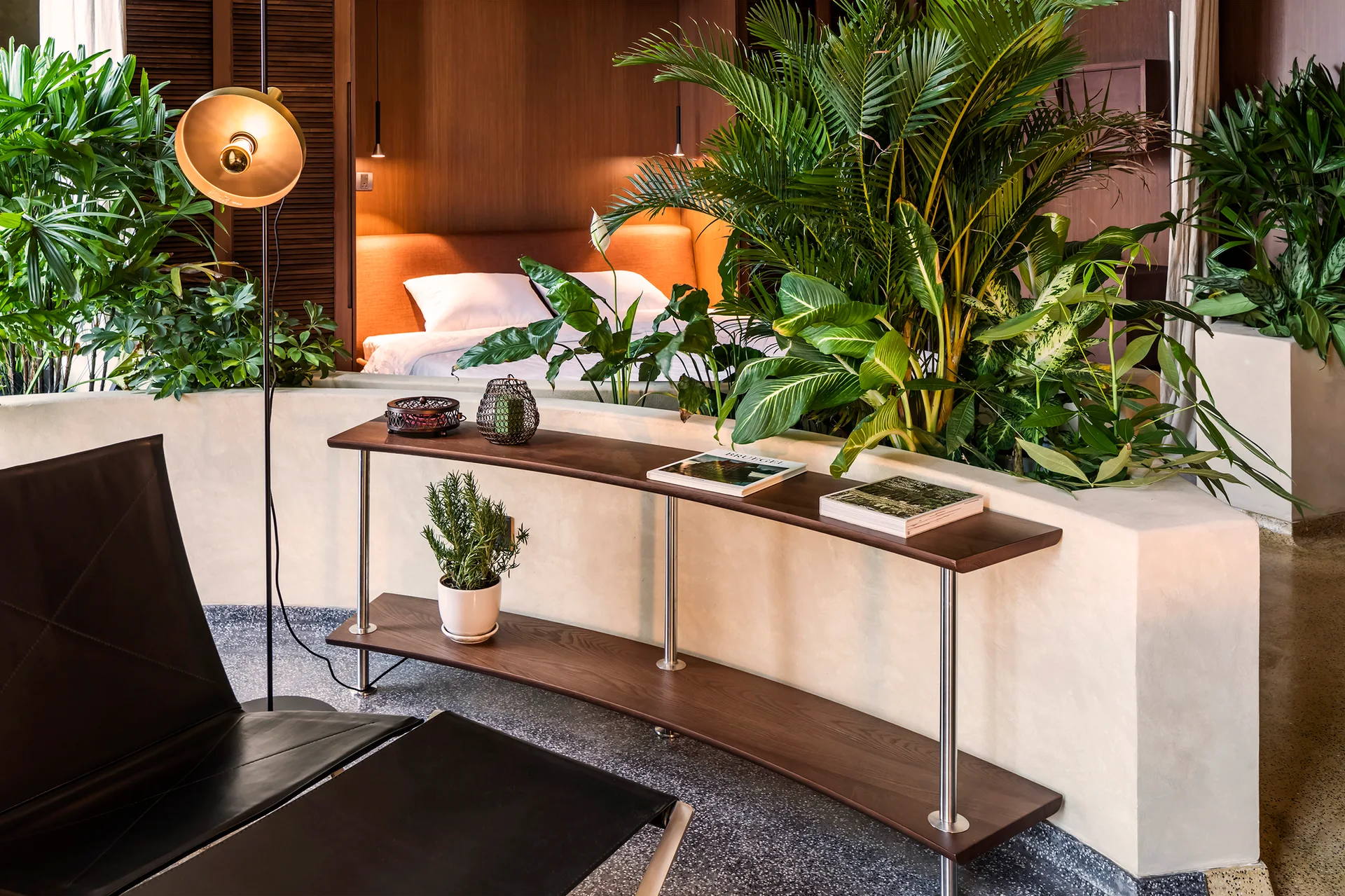 A curved beige plaster wall with integrated tropical plants divides a modern bedroom area from a lounge space with a black leather chair.