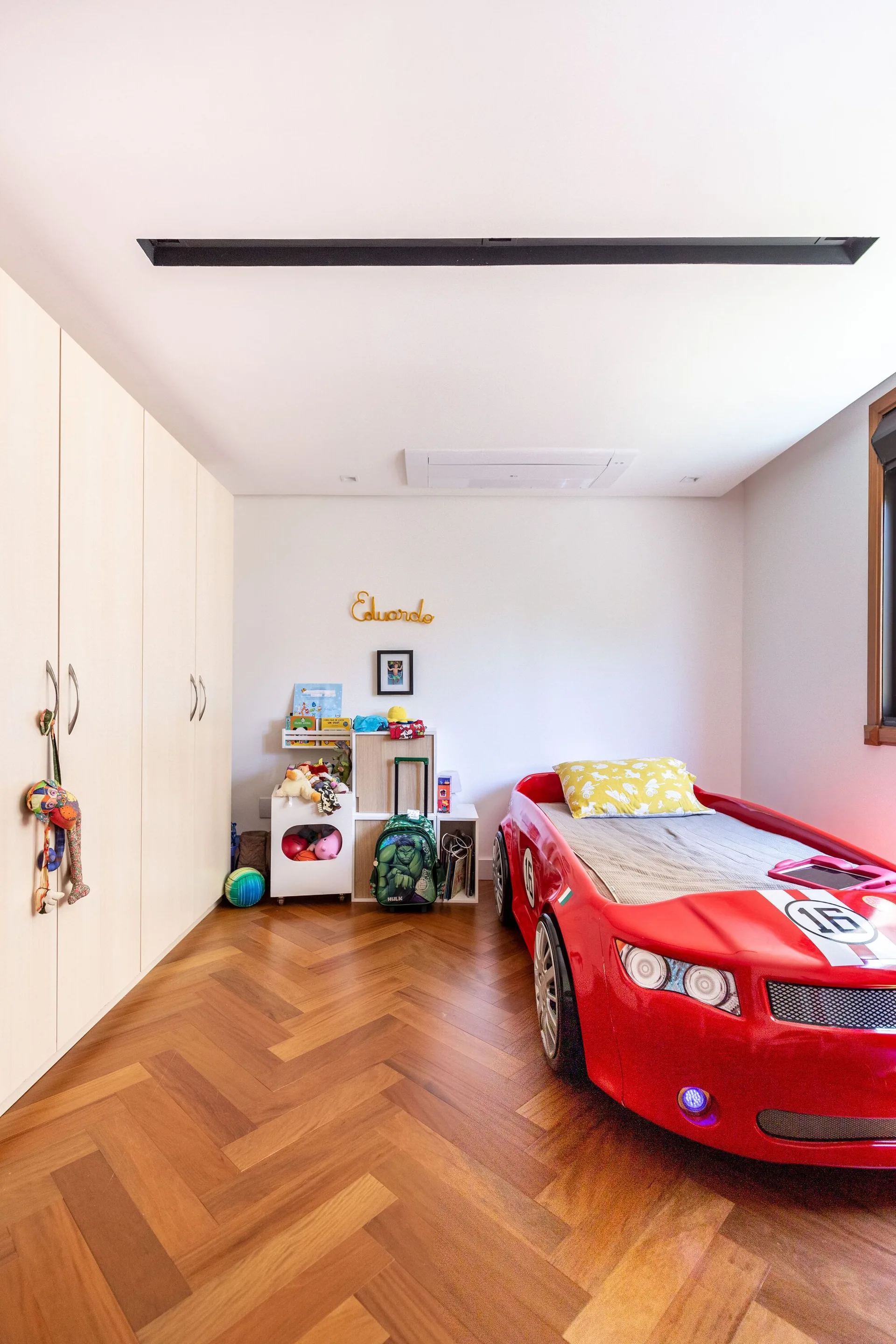 A child's bedroom features a red racing car bed, a light wood floor-to-ceiling wardrobe, and herringbone patterned hardwood flooring.