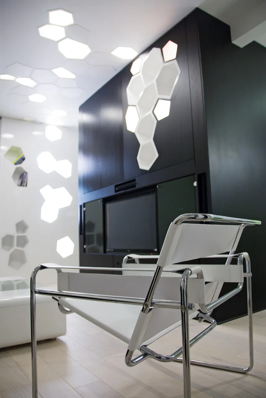 A white leather and chrome armchair in a modern living room with a large black media unit and glowing hexagonal lights on the ceiling and walls.