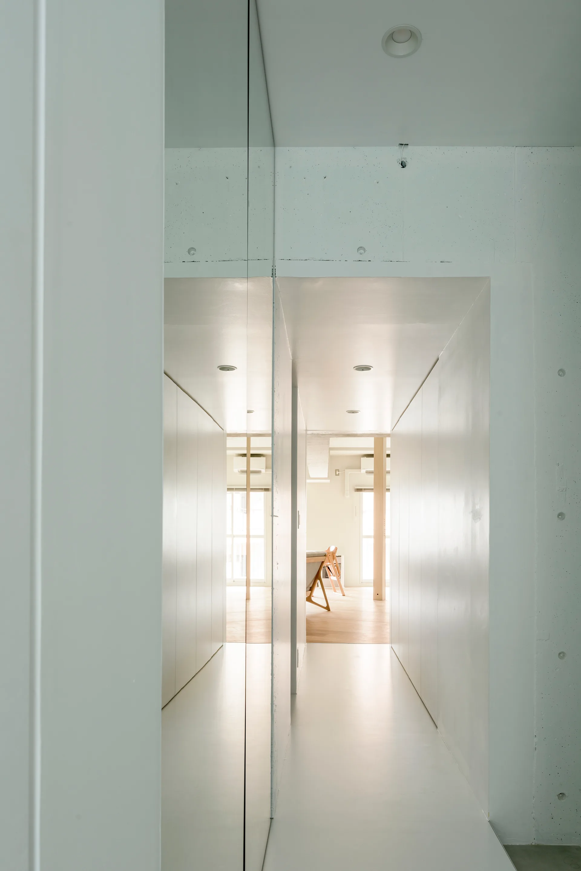 A narrow white hallway with a full-length mirror wall, seamless white cabinets, and recessed lighting leading to a sunlit room.
