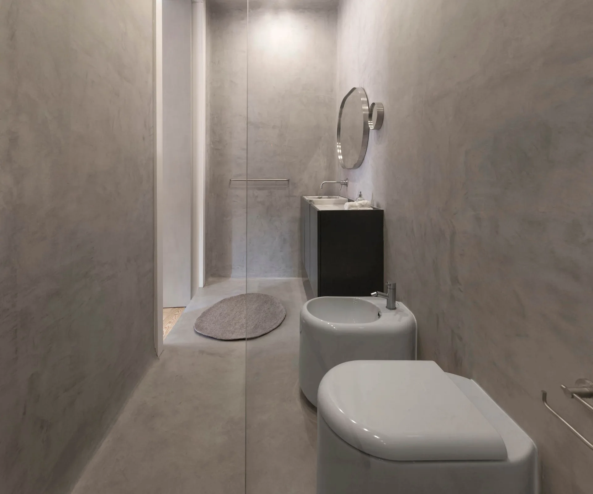 A minimalist bathroom with textured grey concrete walls, a dark floating vanity, a round wall mirror, and modern white toilet fixtures.