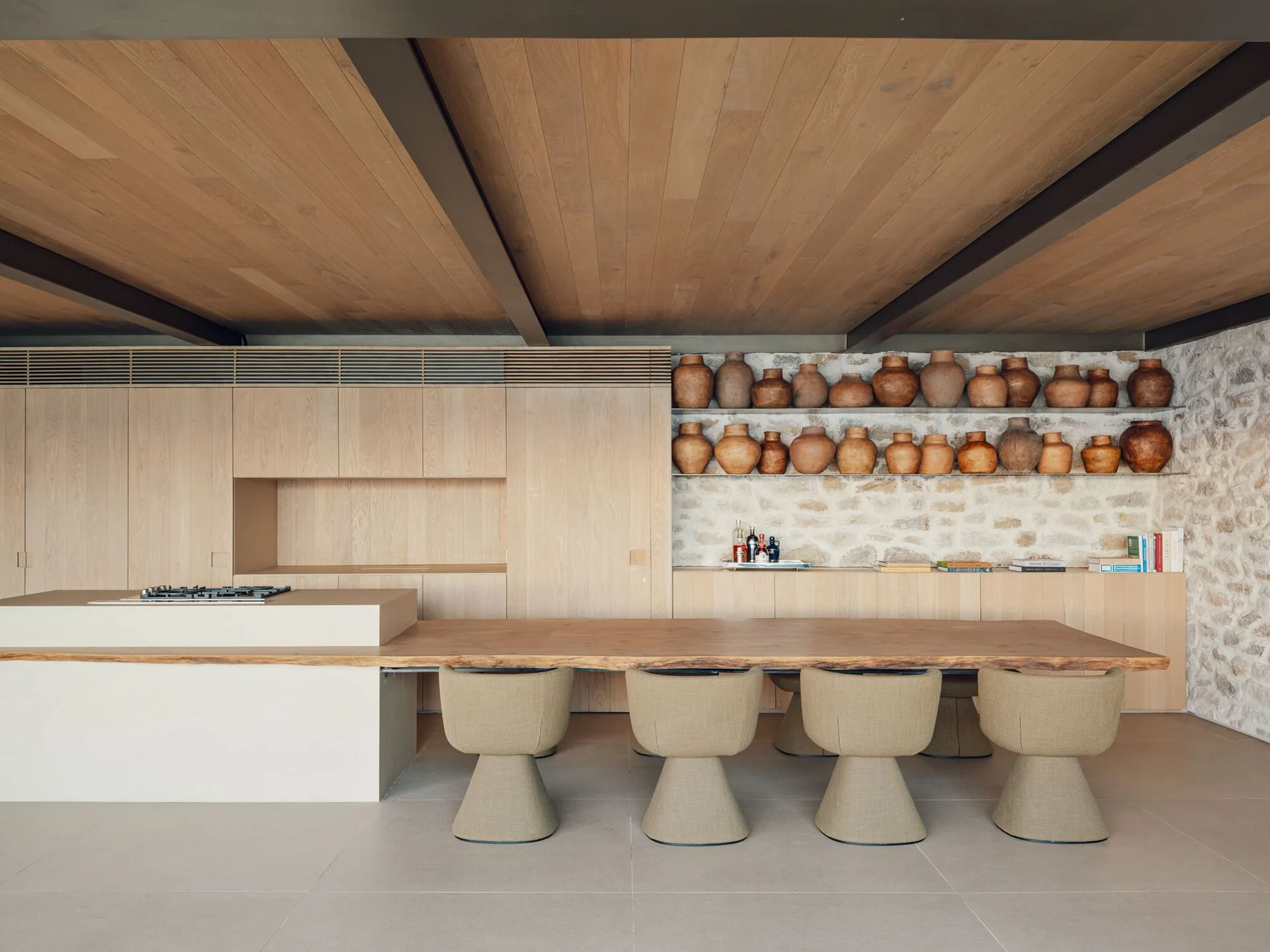 A modern kitchen featuring a long live-edge wood dining table attached to a beige island, backed by light wood cabinets and a stone wall with clay pots.