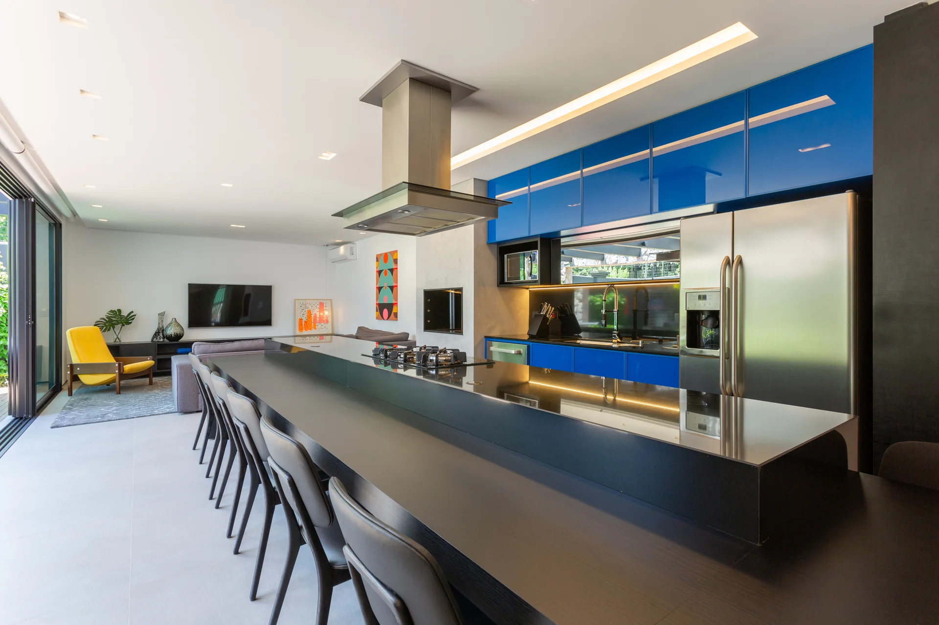 Modern open-plan kitchen featuring glossy blue cabinets, a long black island with bar stools, and a stainless steel refrigerator.