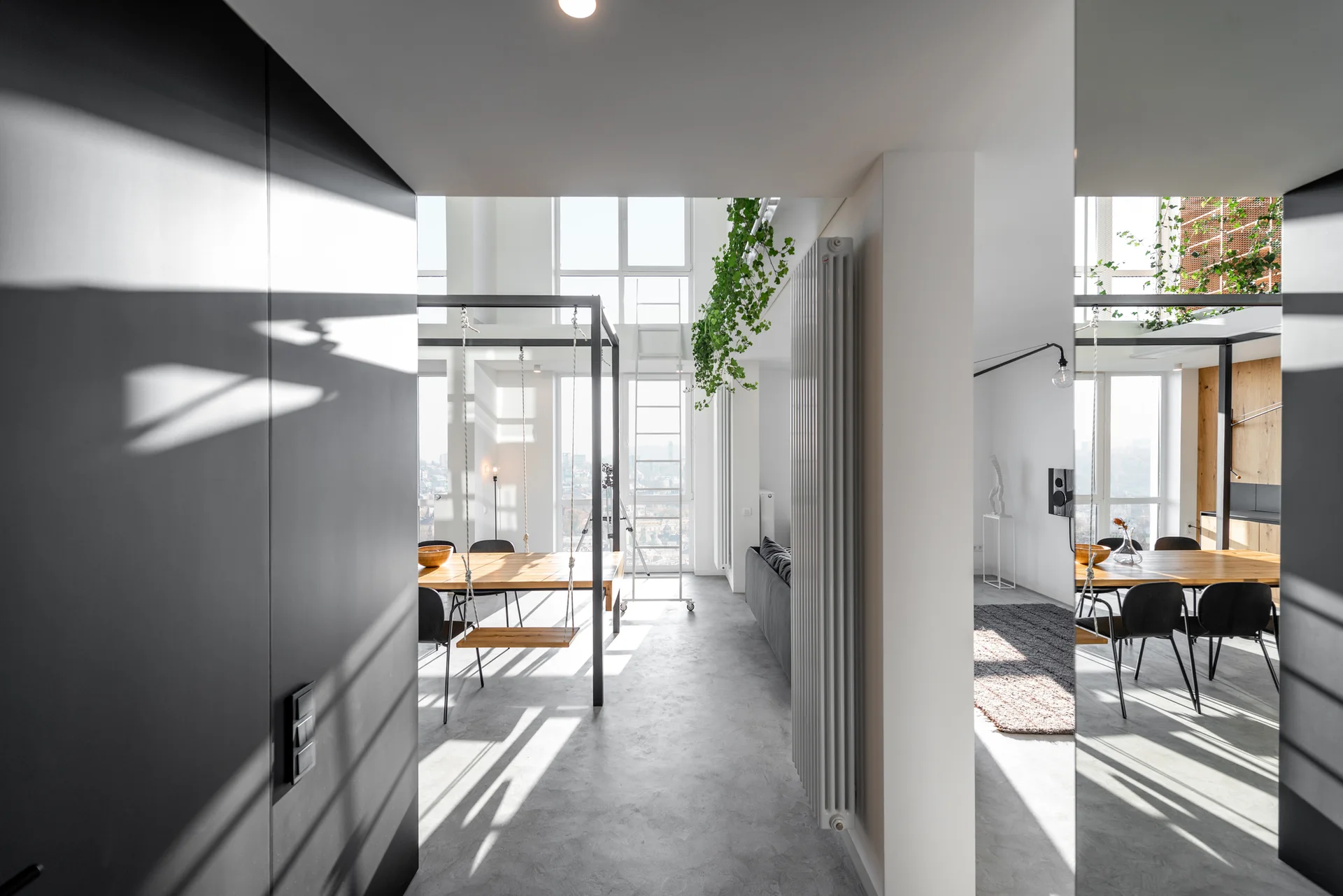 A modern open-plan dining space featuring a wooden table with suspended swing seats, grey concrete floors, and large windows.
