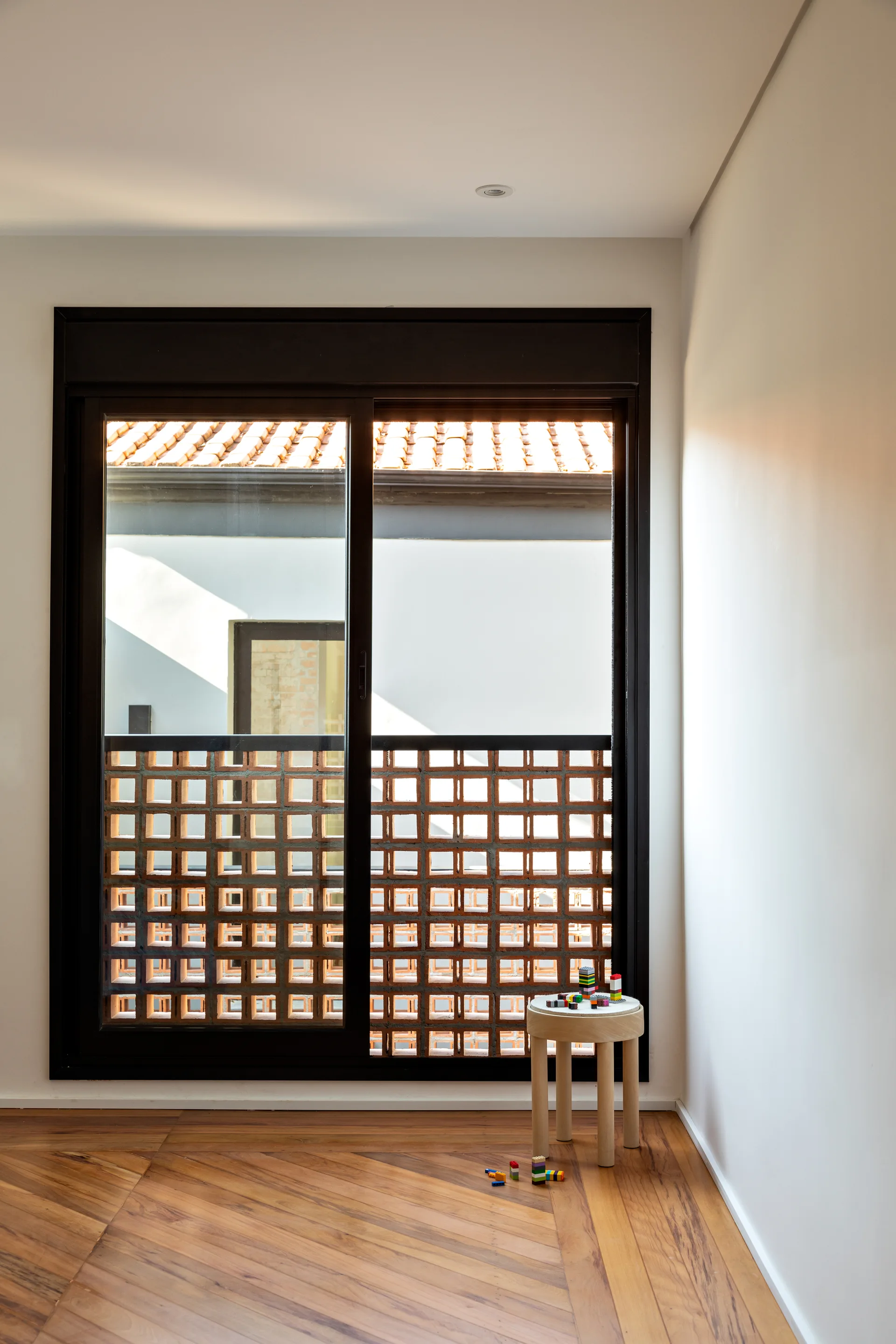A minimalist room with herringbone wood floors features a large sliding window covered by an exterior terracotta breeze block screen.