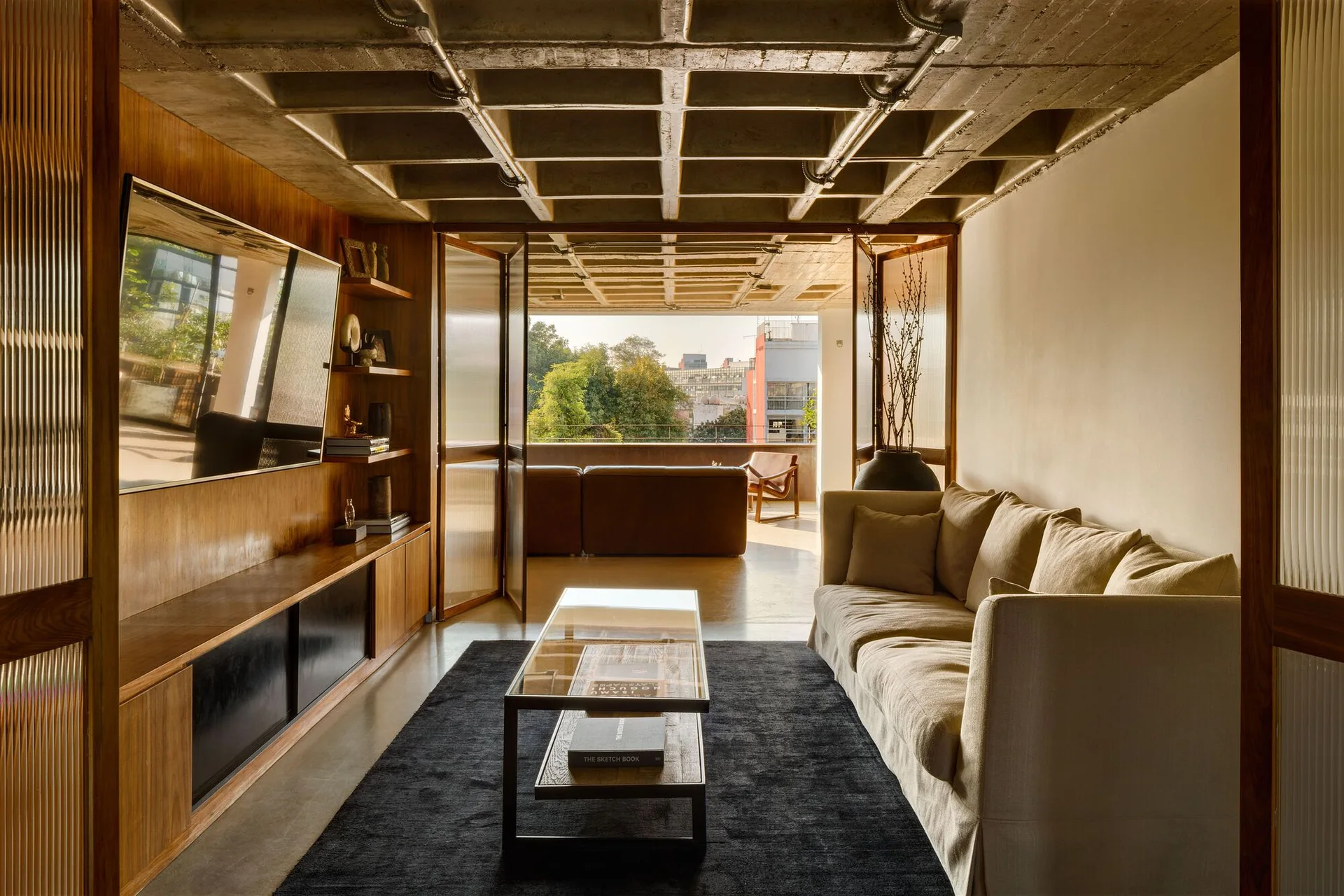 A modern living room features a beige sofa, a glass coffee table on a dark rug, a concrete waffle ceiling, and wood wall shelving.