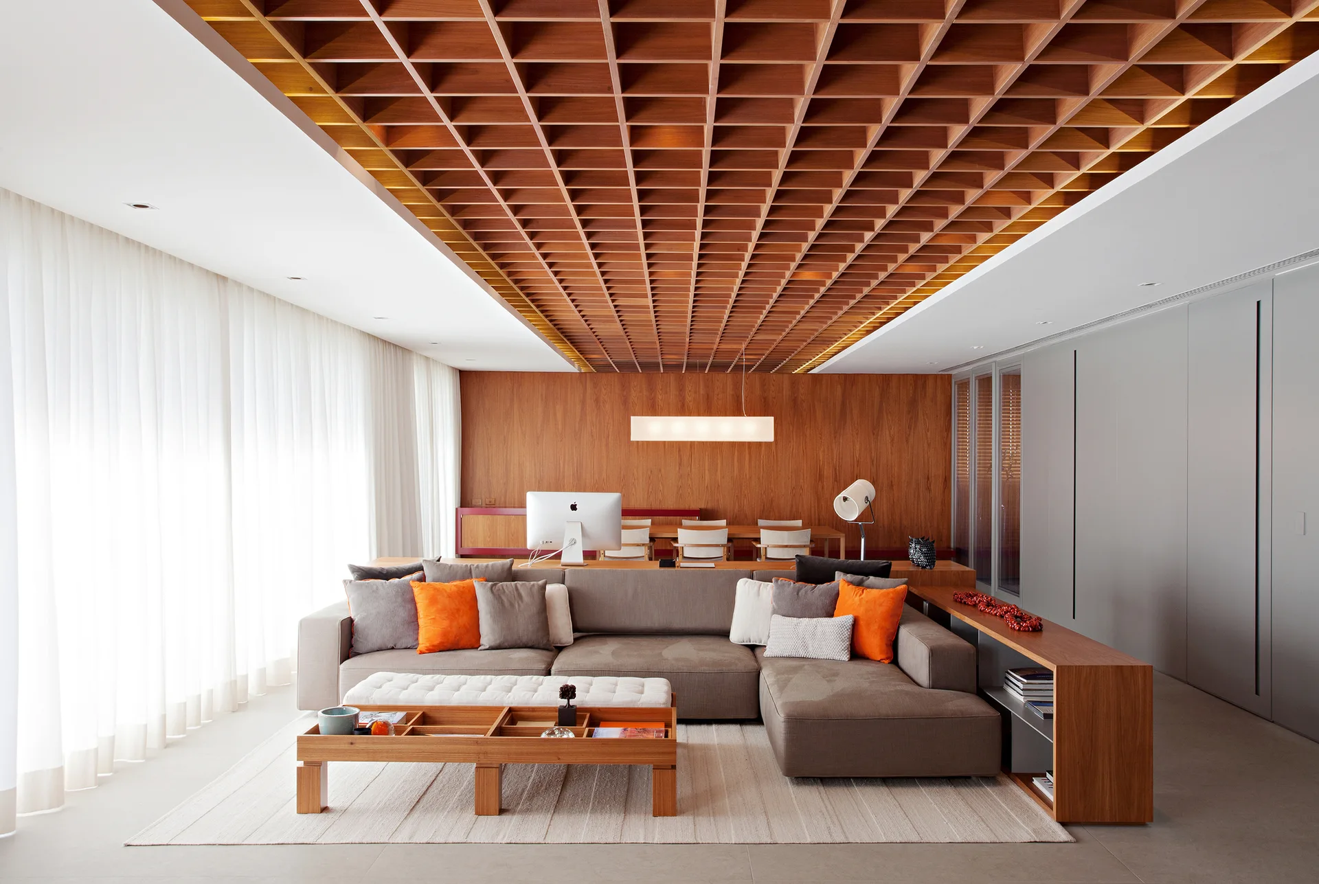 A contemporary living room featuring a wooden coffered ceiling, a grey sectional sofa with orange pillows, and a low wooden coffee table.