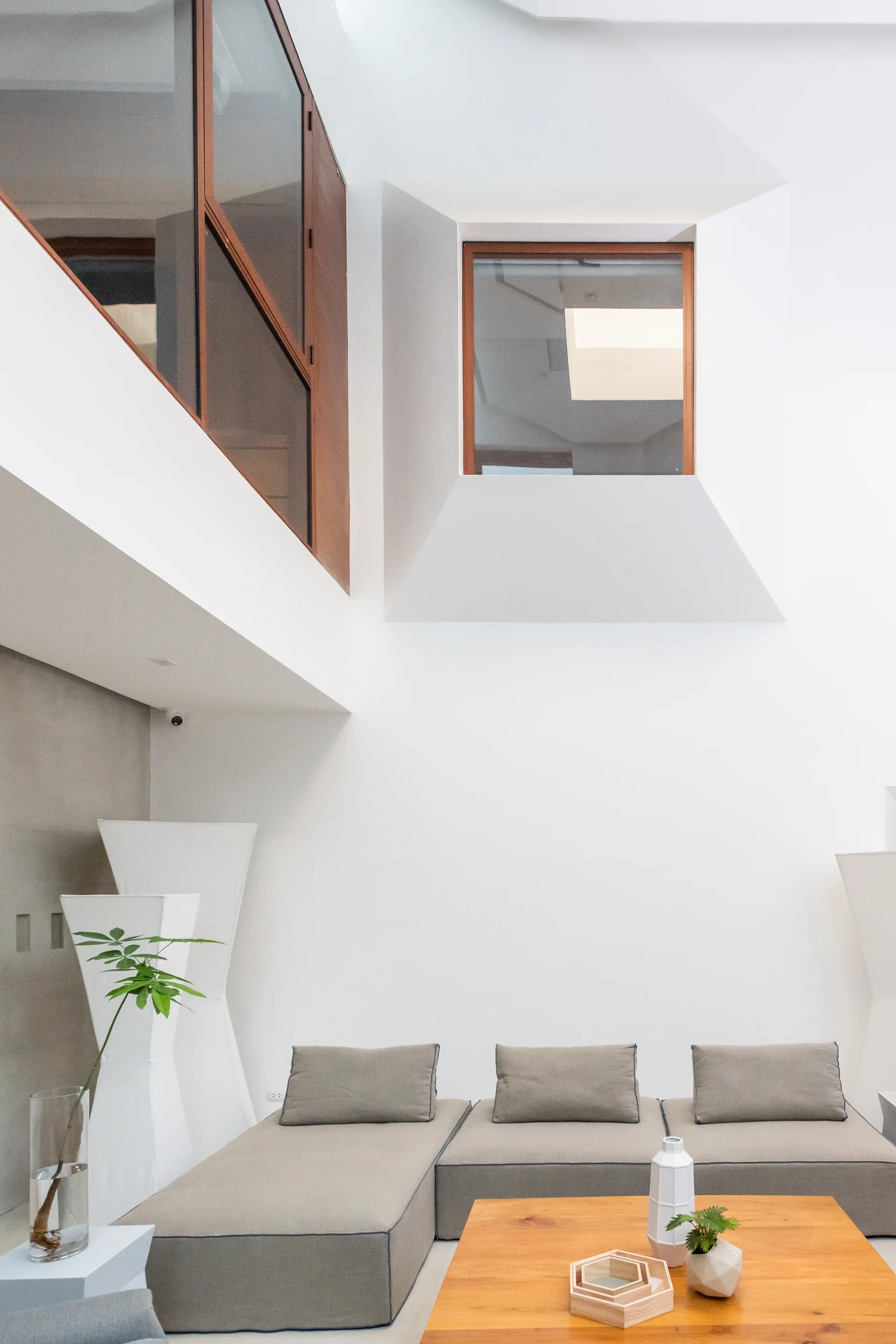 A minimalist living room features a low-profile beige sectional sofa, a wooden coffee table, and wood-framed interior windows on high white walls.