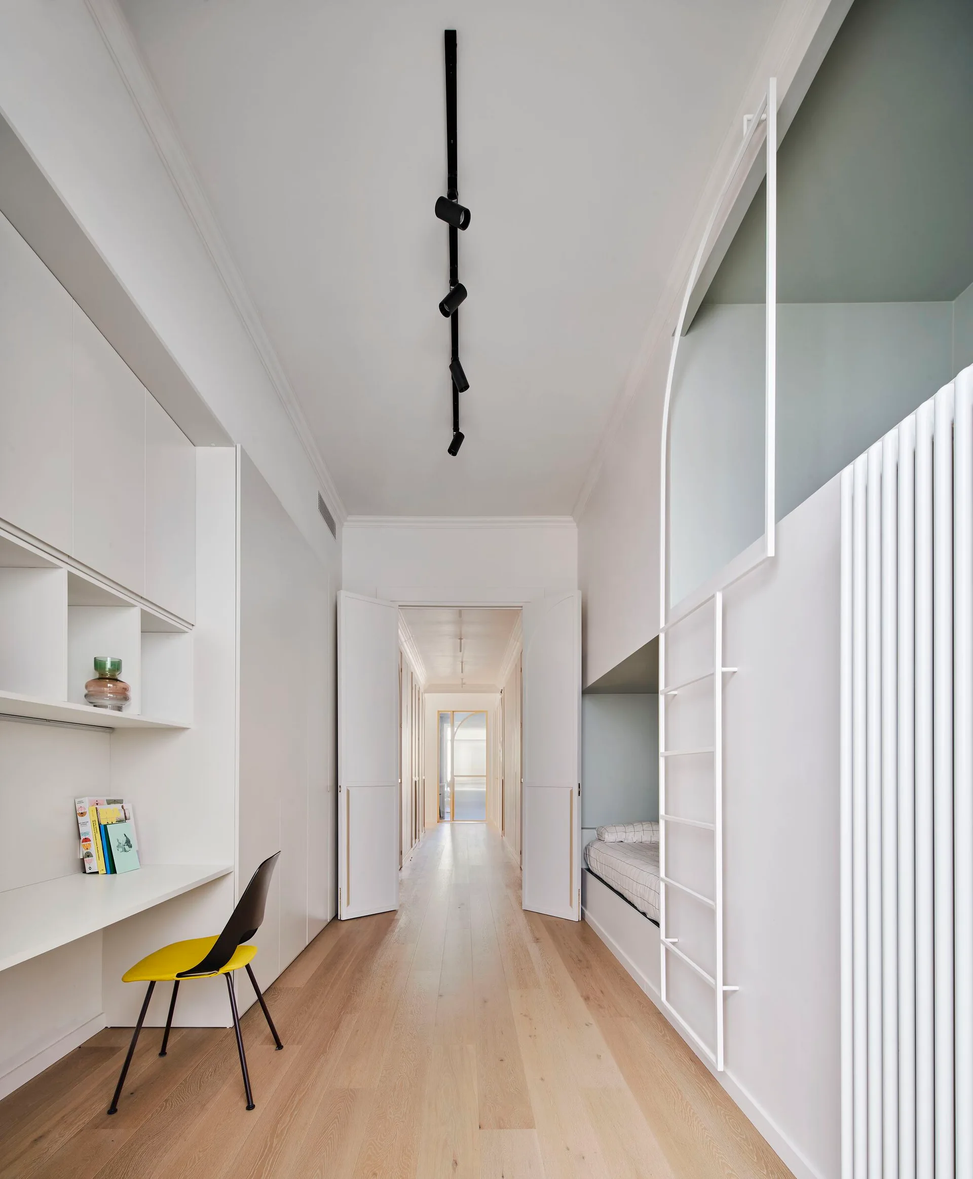 A minimalist white bedroom featuring a built-in desk with a yellow chair, a recessed sleeping nook, a loft ladder, and light wood floors.