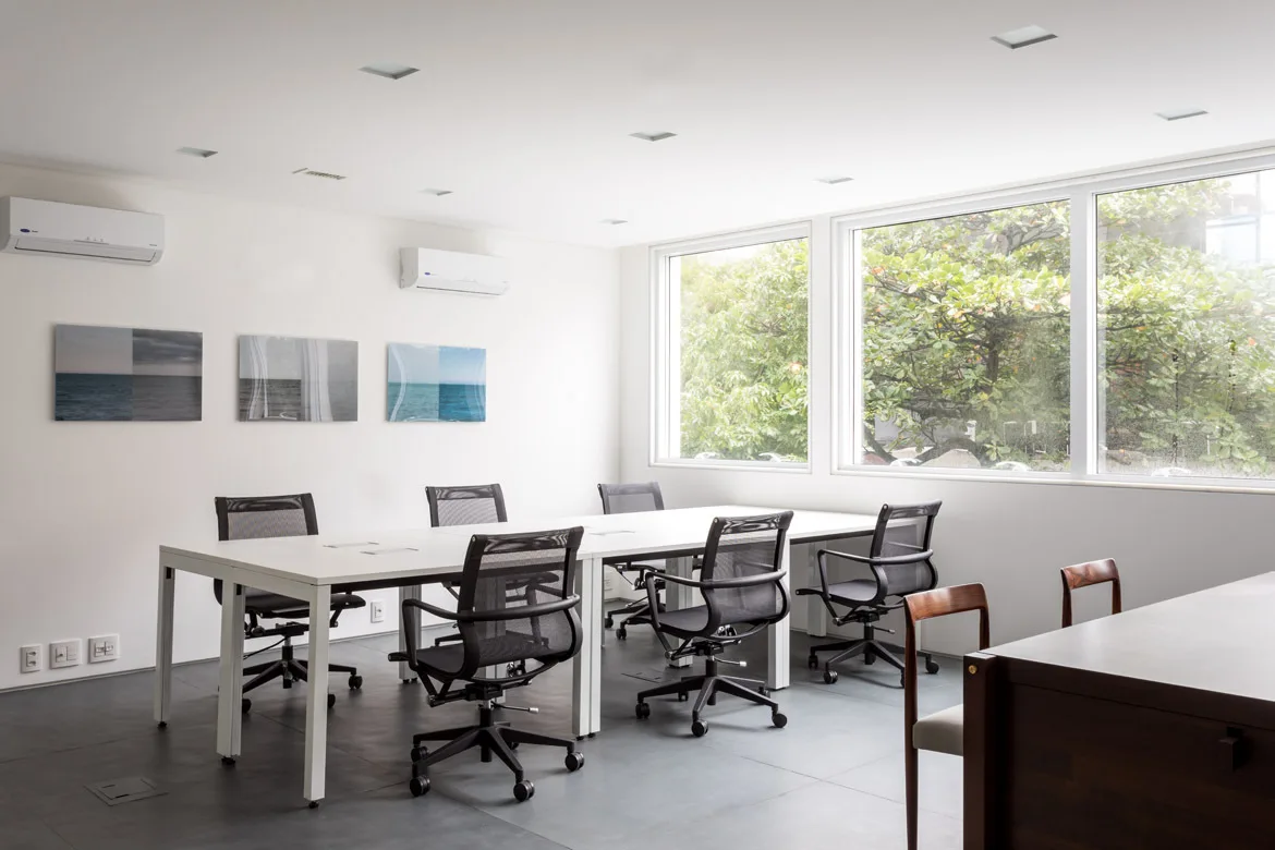 A bright minimalist office conference room featuring a long white table, black mesh rolling chairs, and large windows looking out to leafy green trees.