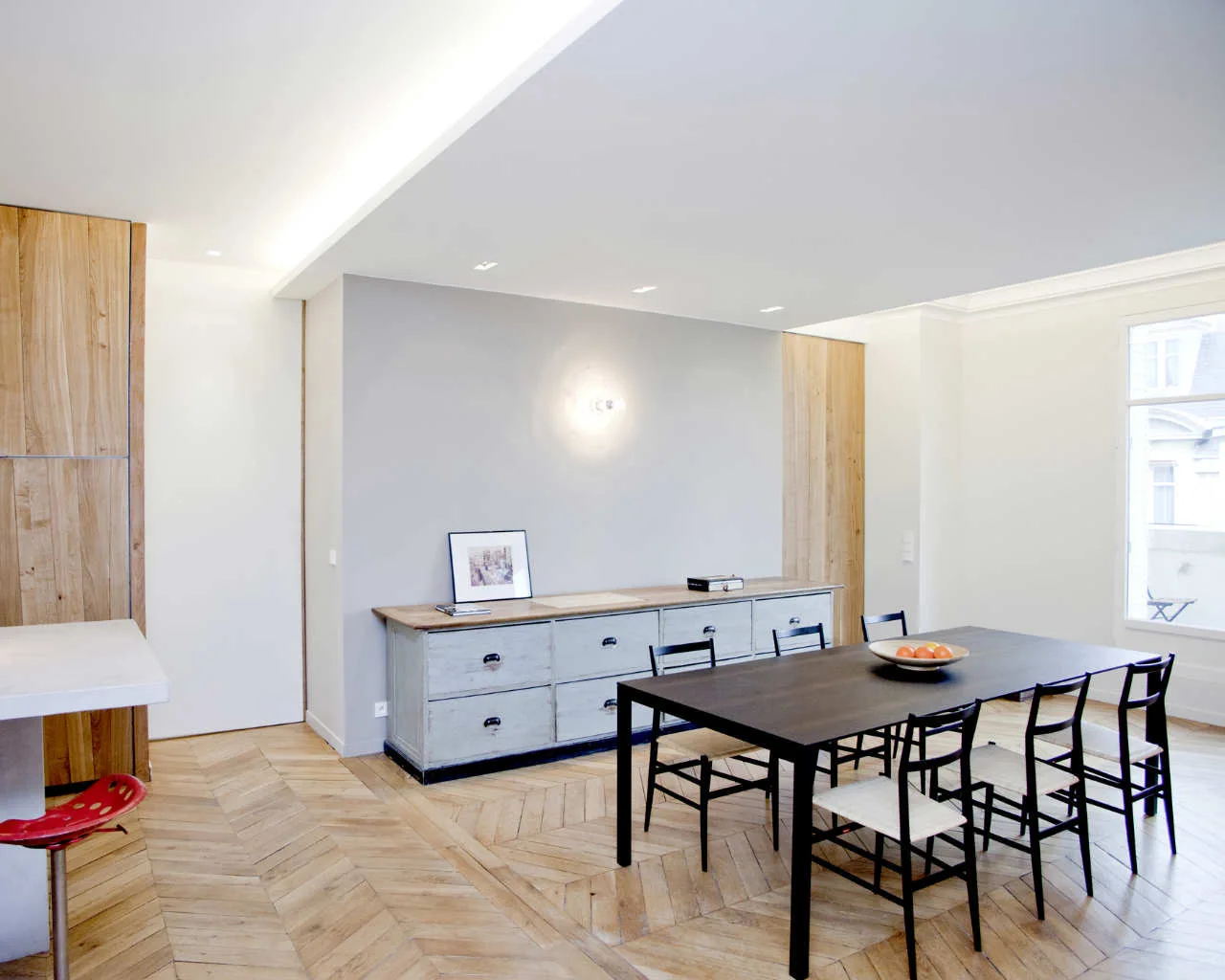 A modern dining room features a dark wood table with black chairs, a light grey sideboard, and light brown herringbone wood flooring.