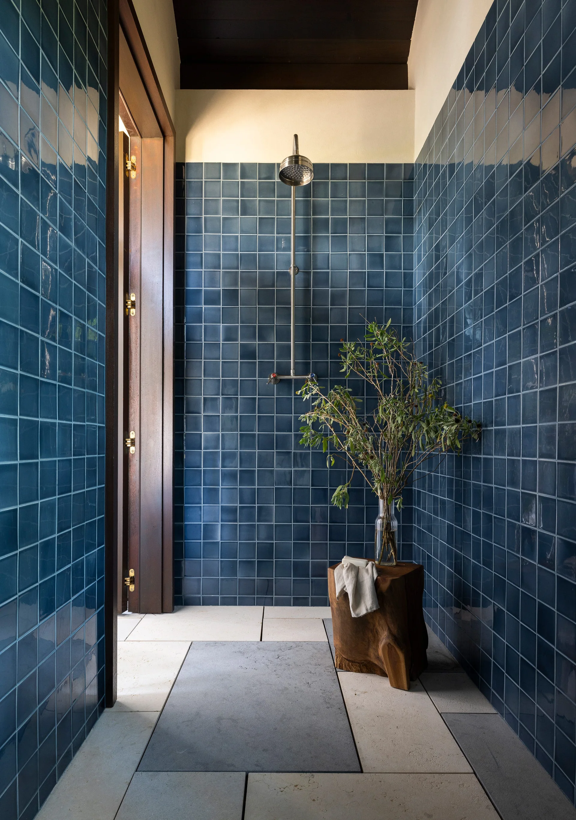 A walk-in shower features glossy deep blue square wall tiles, large off-white stone floor tiles, a natural wood stool with a vase, and a silver shower head.