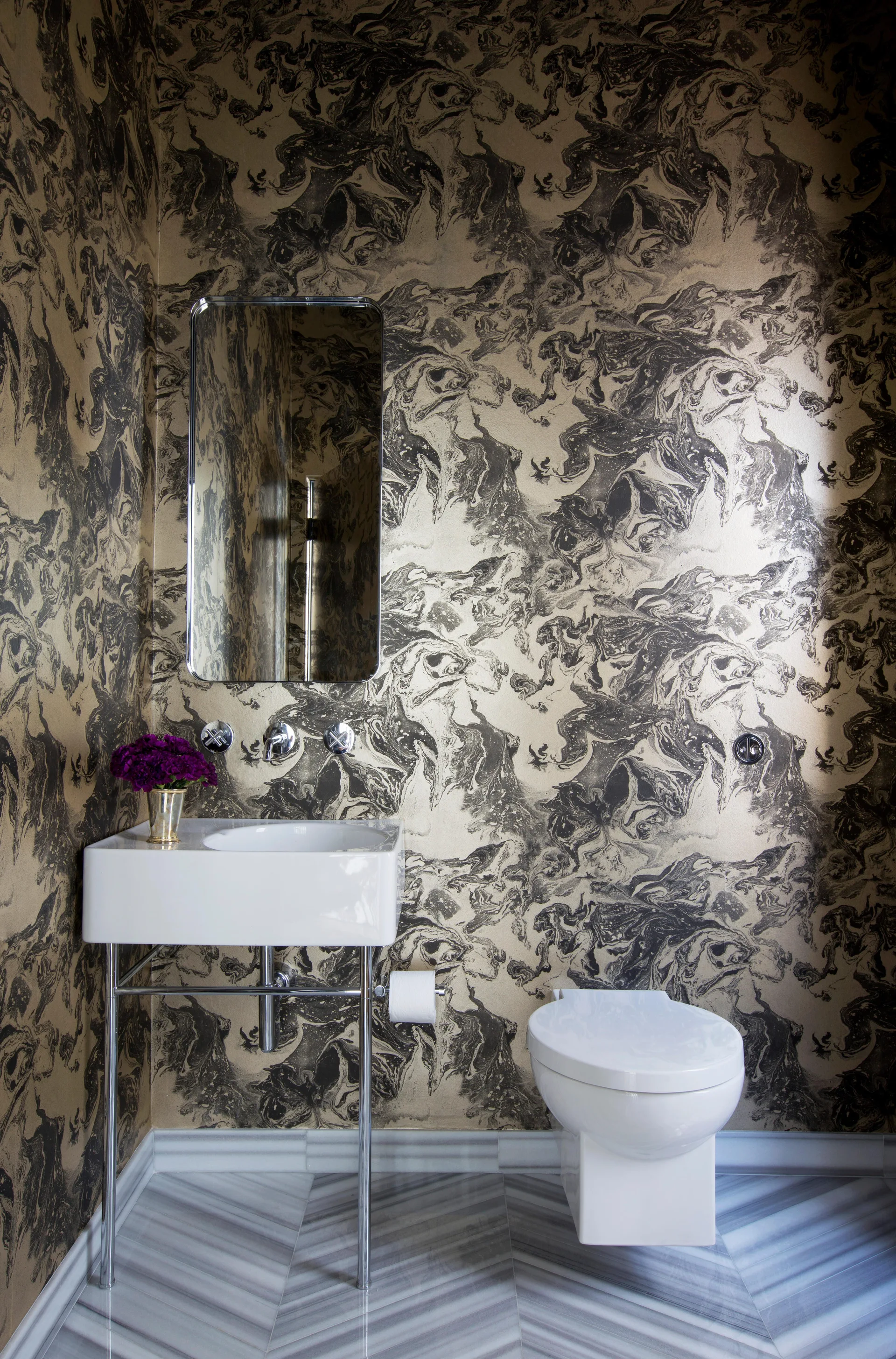 A powder room features dark marbleized wallpaper, a white console sink with metal legs, a rectangular mirror, and chevron marble tile flooring.