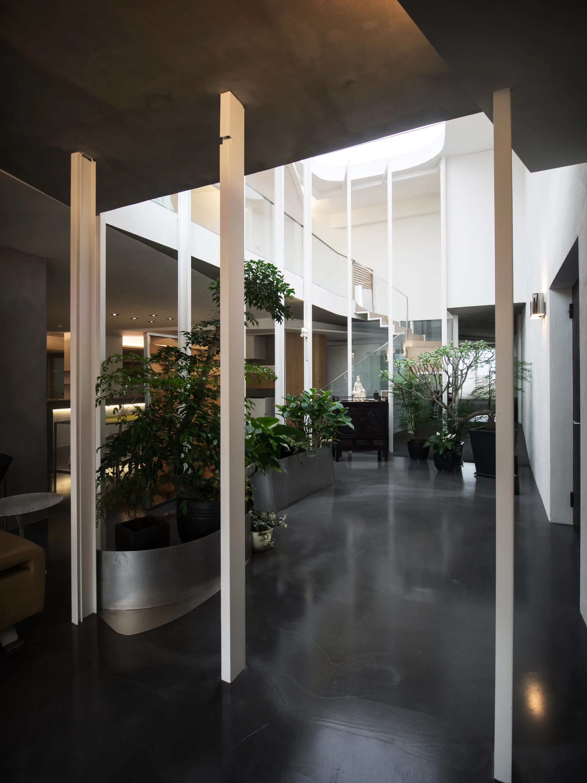 A minimalist indoor atrium featuring vertical white columns, a glossy dark floor, curved metal planters with trees, and a glass-railed upper walkway.