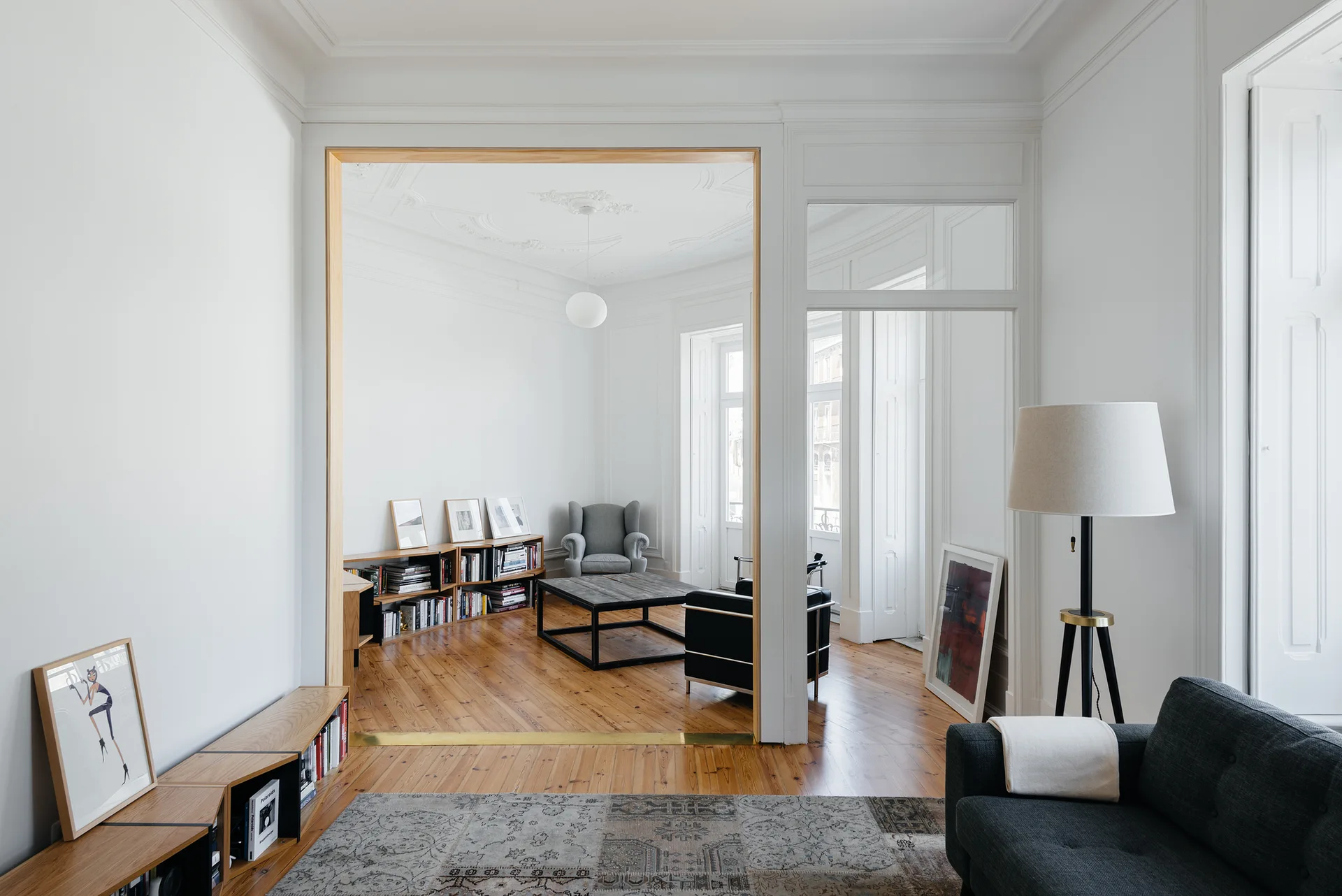 A bright living room with white walls and wood floors features a large natural wood cased opening leading into an adjacent seating area.