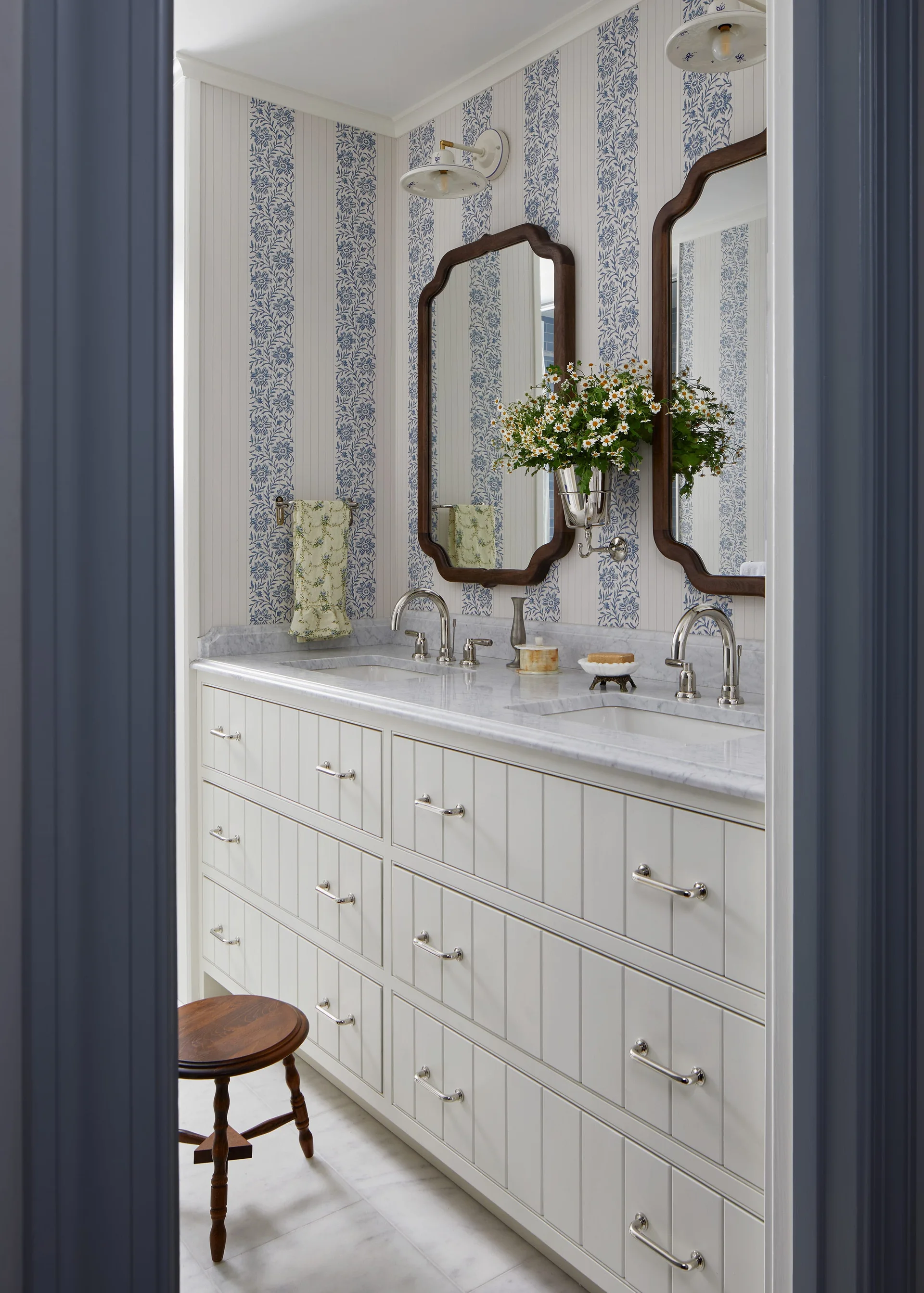 A bathroom vanity with a marble countertop, white paneled drawers, two wood-framed scalloped mirrors, and blue and white striped floral wallpaper.