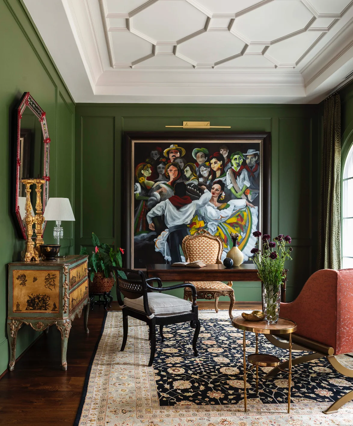 A home office featuring forest green paneled walls, a white coffered ceiling, a dark wood writing desk, and a large colorful painting.