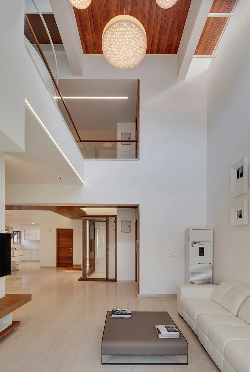 A double-height living room with white walls, a polished tile floor, a white leather sofa, and a wood-paneled ceiling with orb pendant lights.