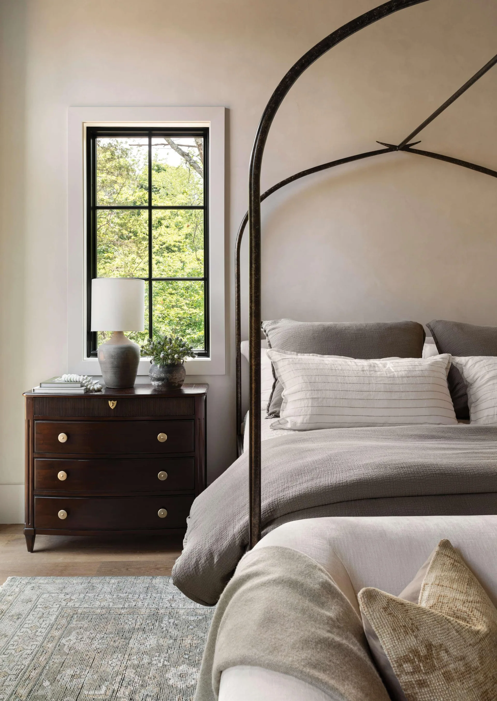 A neutral bedroom features a dark iron four-poster bed and a dark wood chest of drawers next to a black grid window.