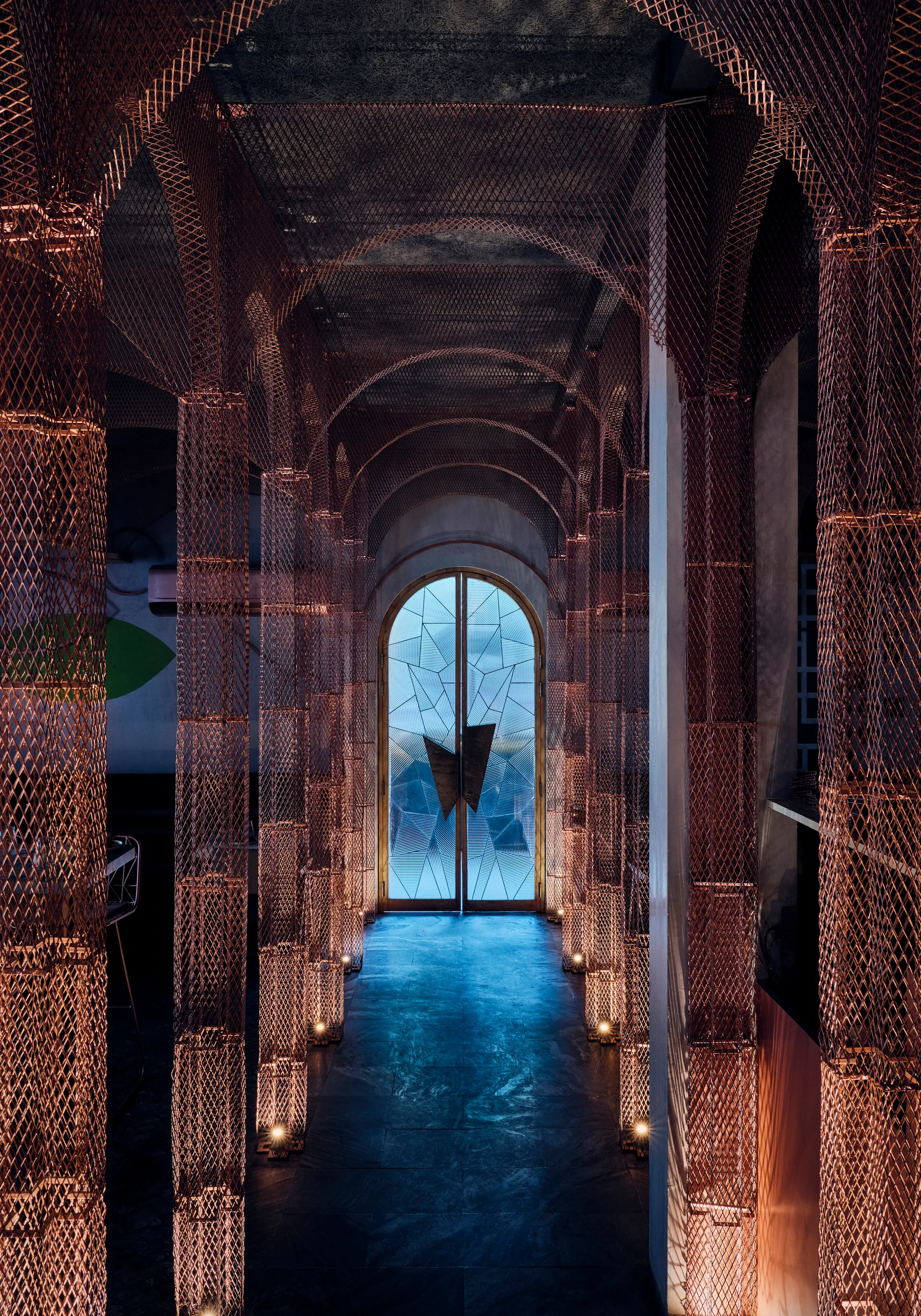 A narrow hallway featuring copper-toned metal mesh columns and a vaulted ceiling leading to a geometric arched glass door.
