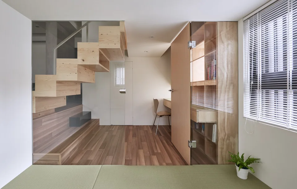 A minimalist room featuring a floating wooden block staircase behind a glass partition, a built-in plywood desk and storage, and light wood flooring.