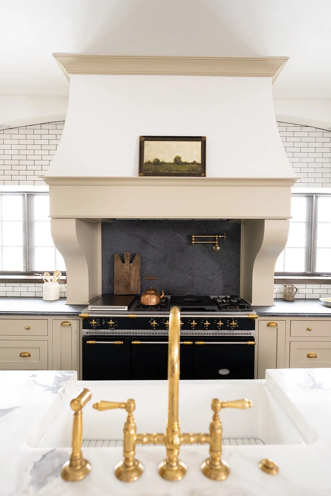 A classic kitchen with a black range under a beige custom vent hood, featuring a brass bridge faucet and farmhouse sink in the foreground.