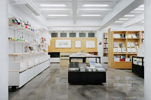 A bright minimalist bookstore interior featuring light wood shelving, white walls, and a polished grey concrete floor.