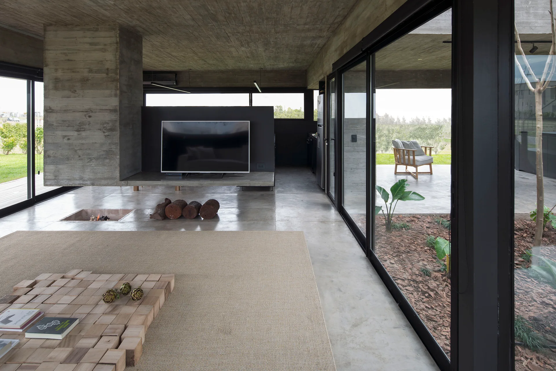 A minimalist living room with raw concrete walls, a sunken floor fireplace, a wooden block coffee table, and large sliding glass doors.