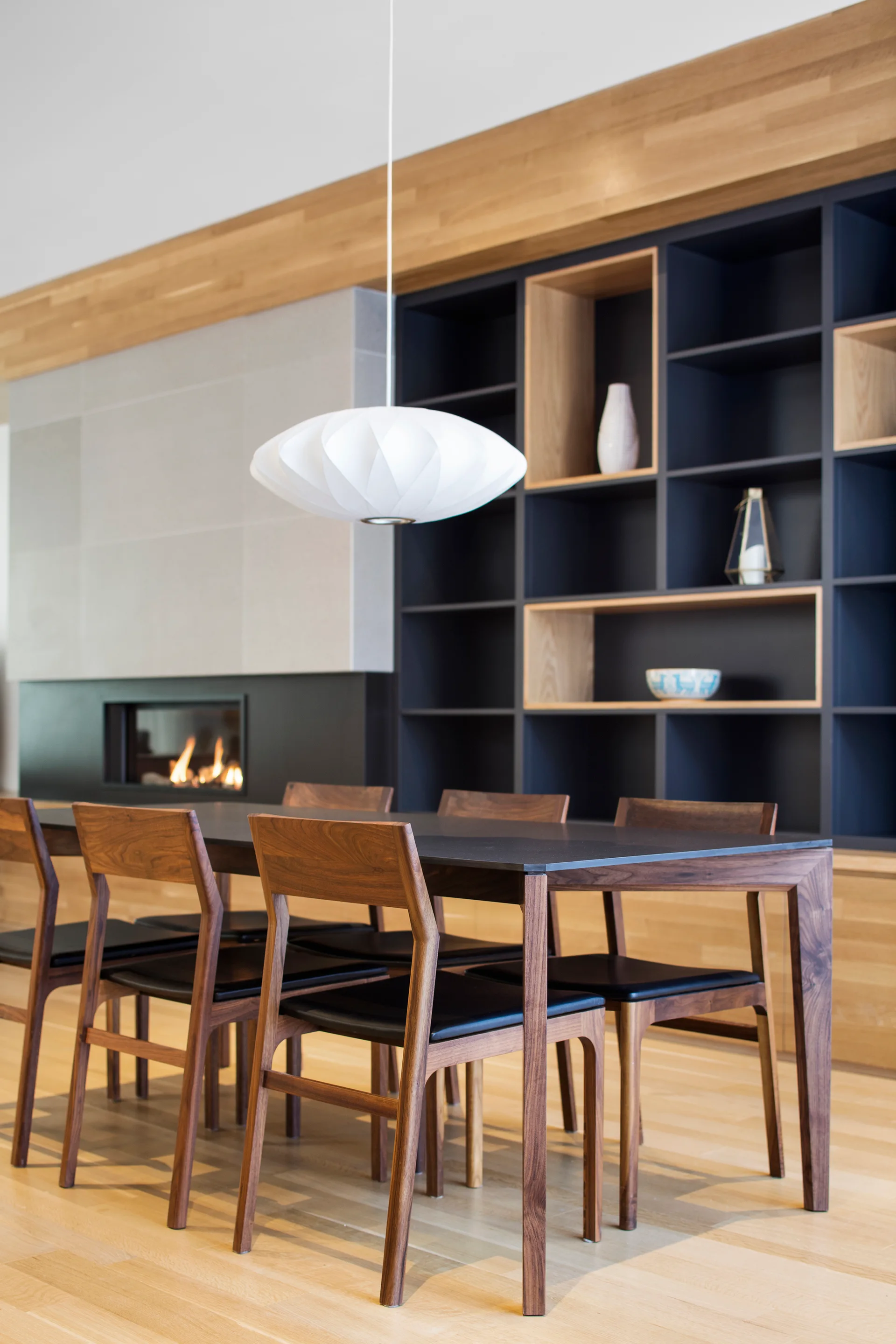 A modern dining room features a dark wood table with matching chairs under a white pendant light, next to a black built-in bookcase and a fireplace.
