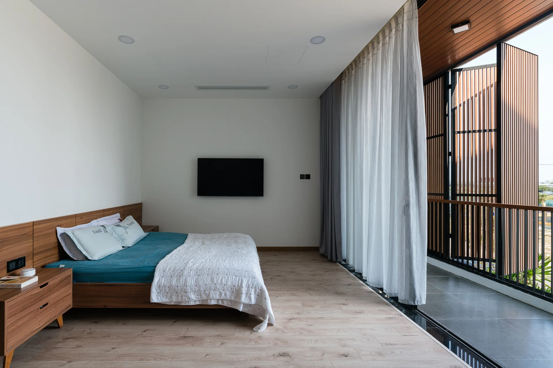A minimalist bedroom with a wood platform bed, an integrated headboard panel, and sheer white curtains opening onto a balcony with slatted wood screens.